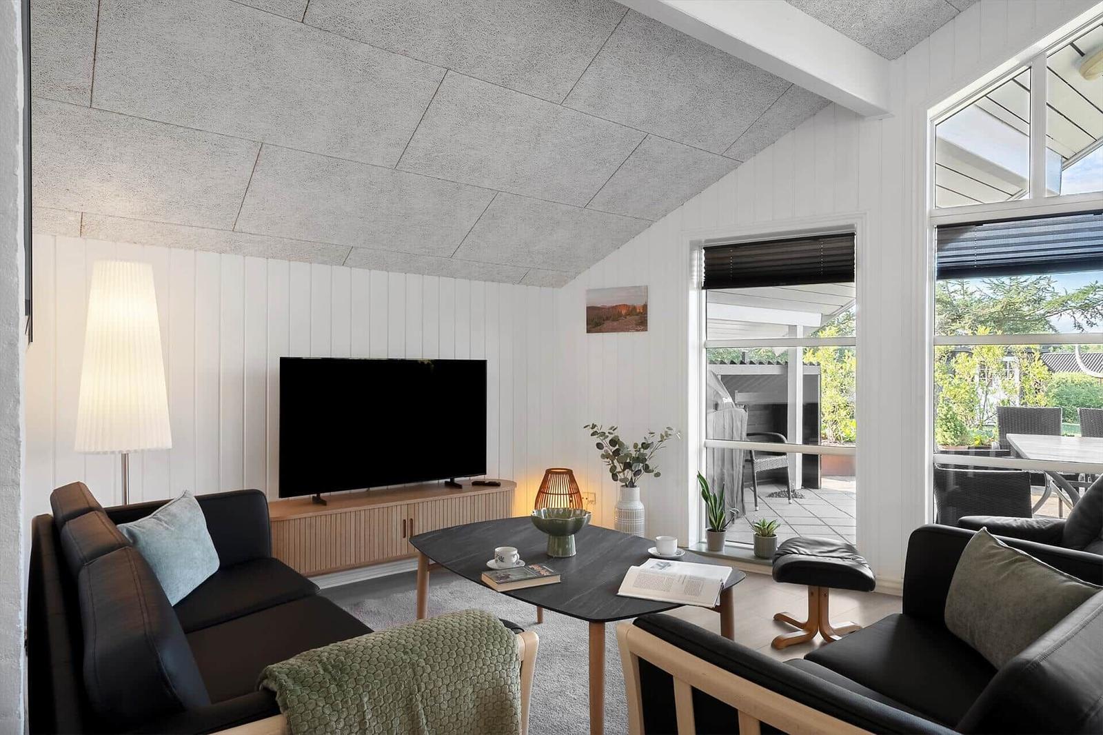 Wohnzimmer mit Sofa, Fernseher und Tisch. Durchgehende Fenster zur Terrasse.