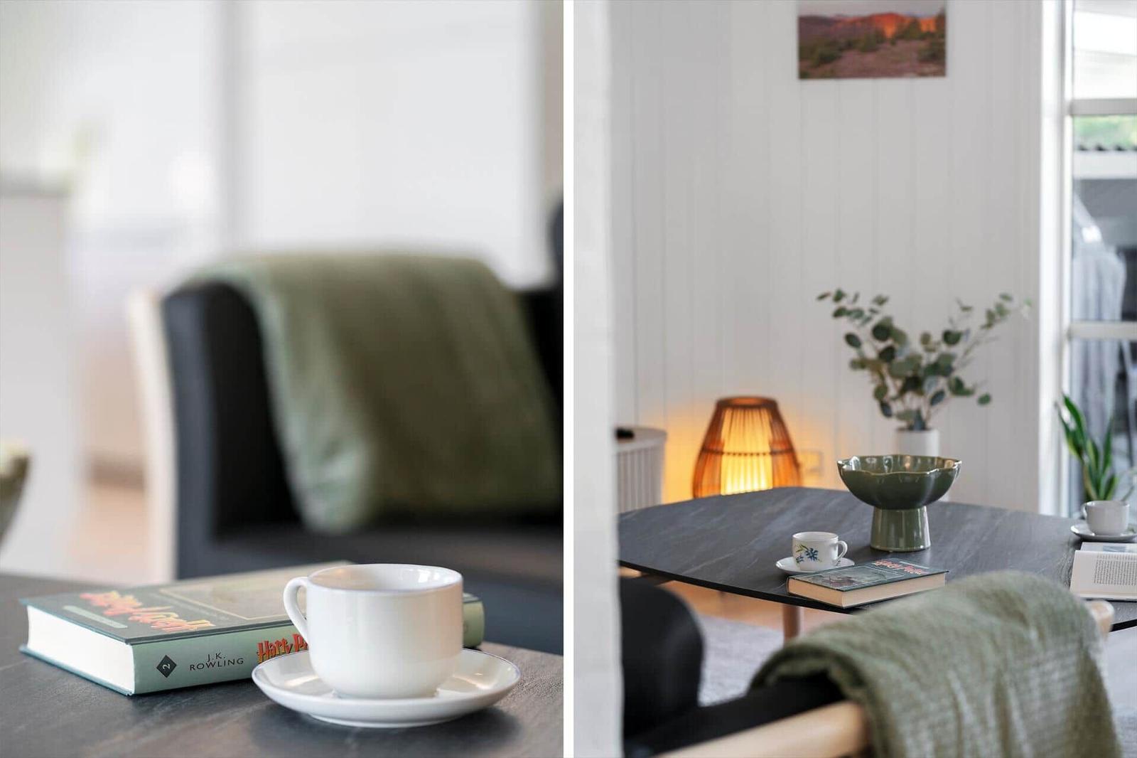 Tisch mit Tasse, Buch und Pflanze. Hintergrund: Sofa mit Decke und Wandbild.