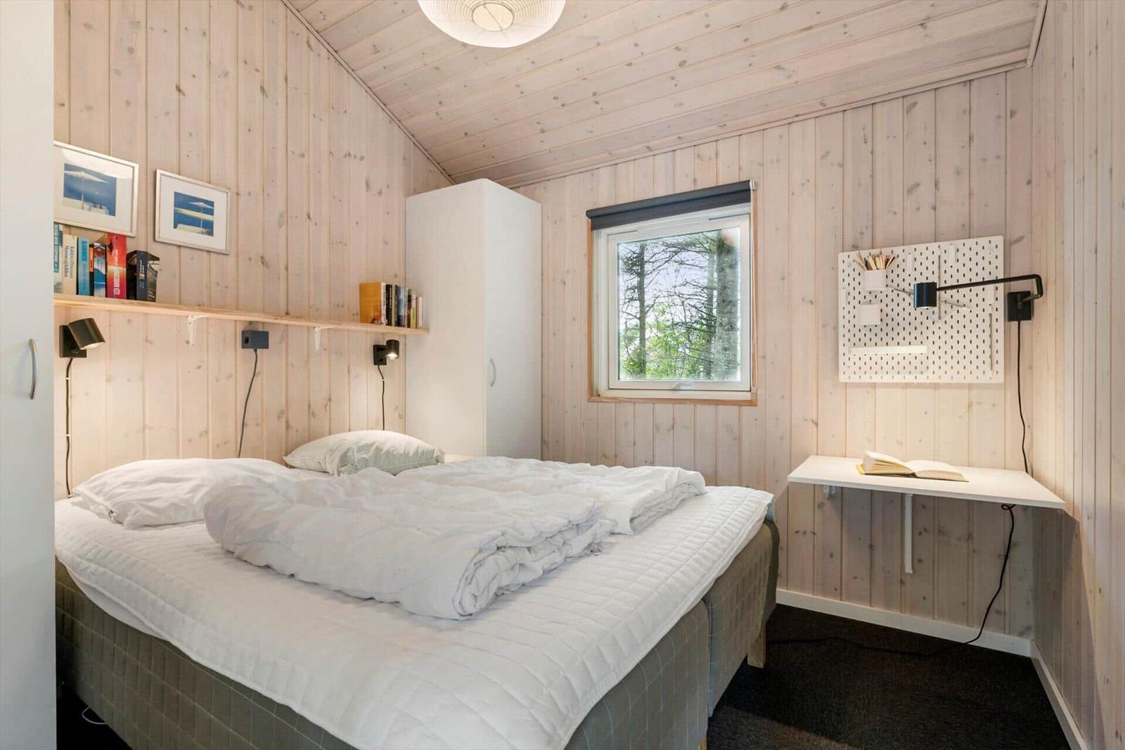Schlafzimmer mit Bett, Schreibtisch und Fenster. Holzwand und weiße Möbel.