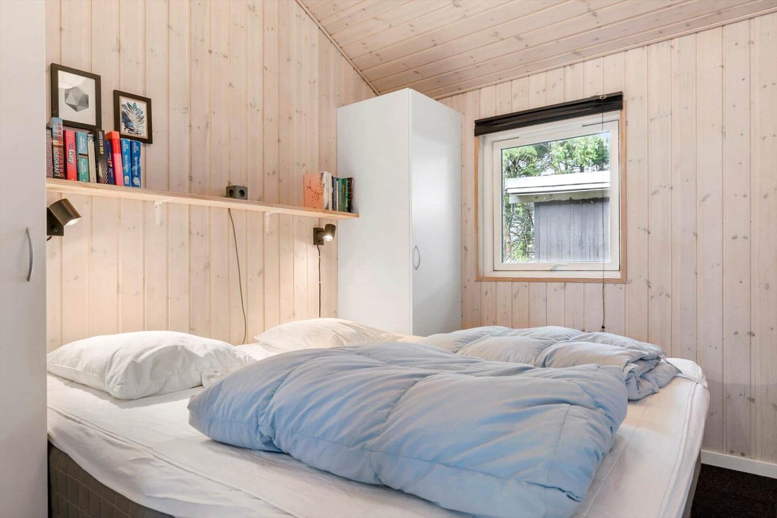 Schlafzimmer mit Bett, Fenster und weißen Schränken an Holzwänden.