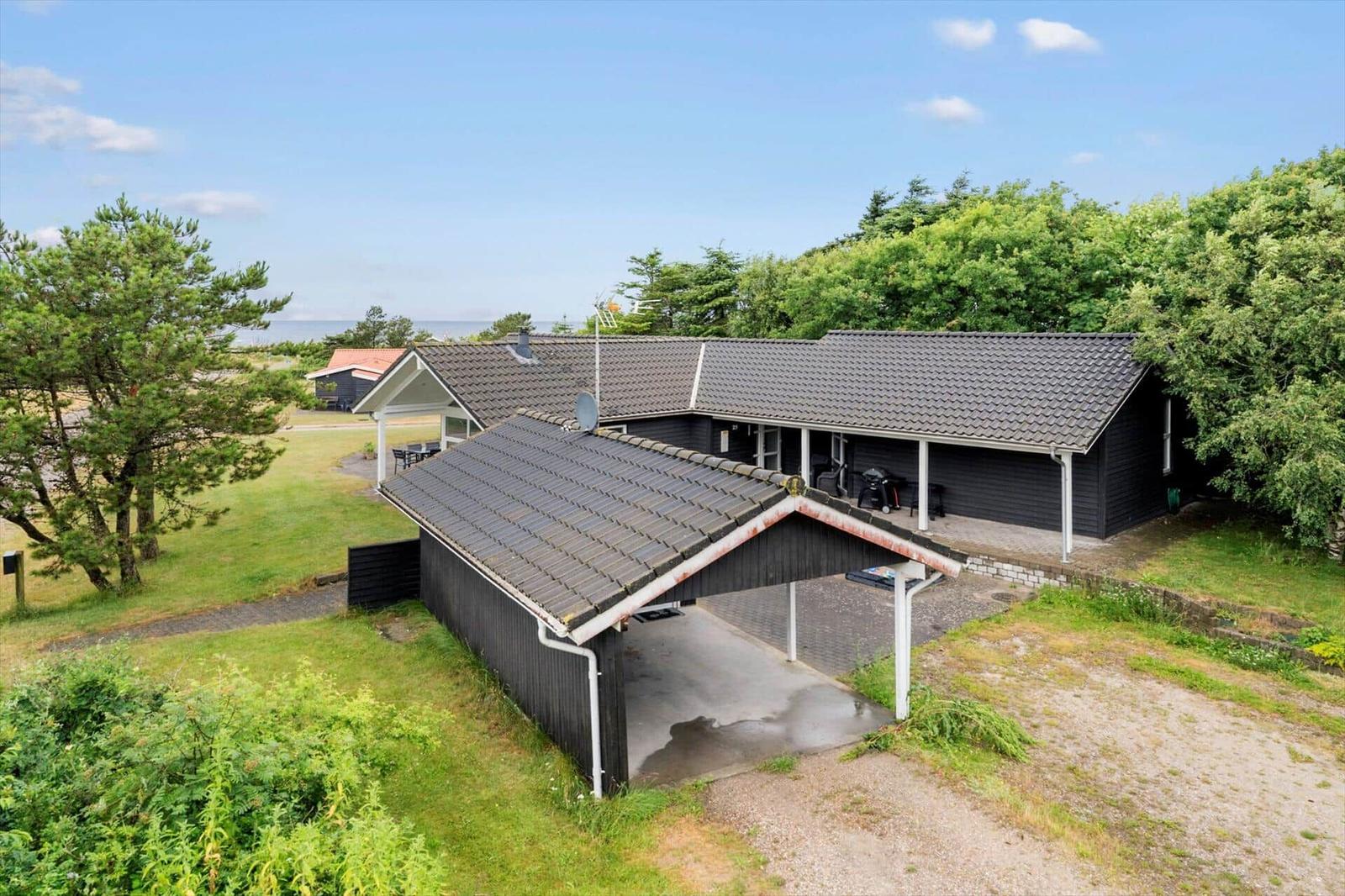 Ein dunkles Haus mit Garage und Terrasse, umgeben von Grünflächen und Bäumen. Der Blick geht zum Meer.