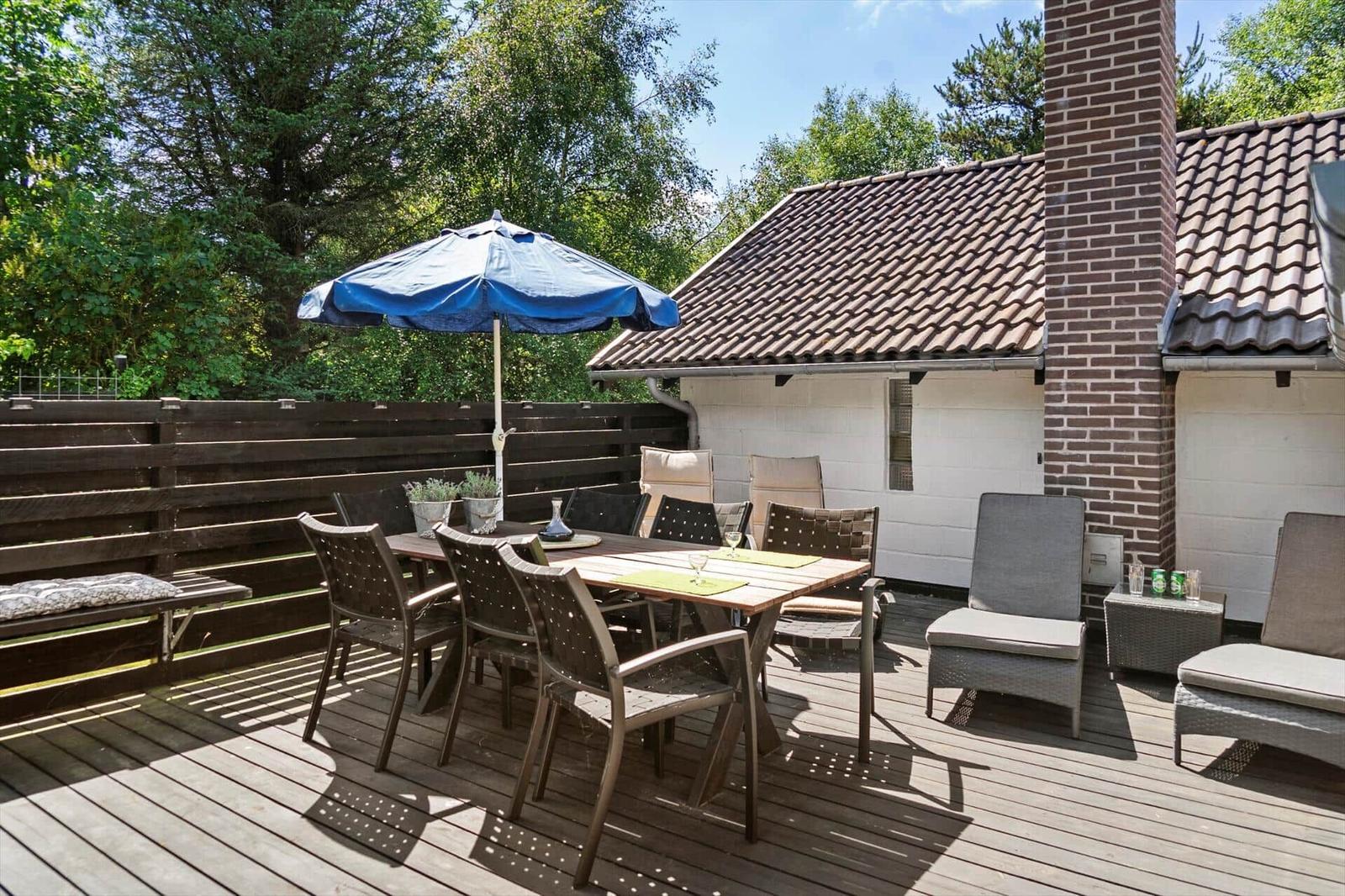 Terrasse mit Tisch, Stühlen und blauem Sonnenschirm. Hintergrund: Haus mit Ziegeldach und Holzgeländer.
