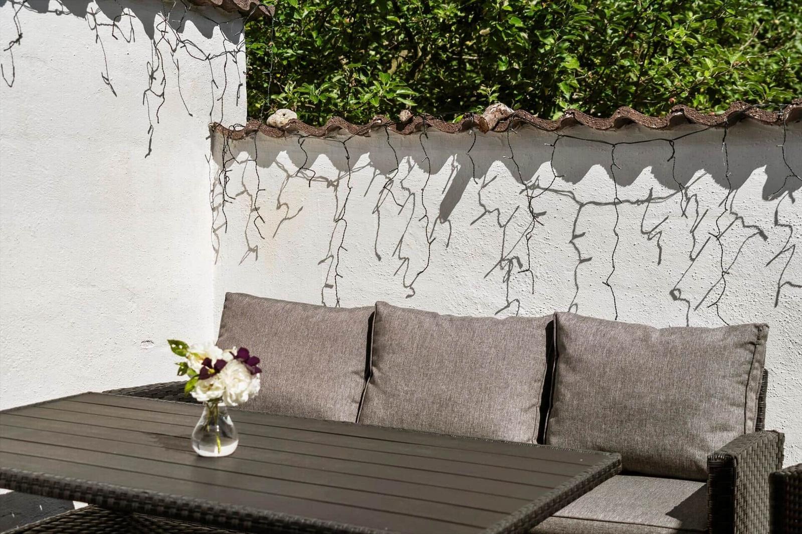 Außenbereich mit Sitzbank, Tisch und Blumenarrangement vor weißer Wand mit Pflanzen.