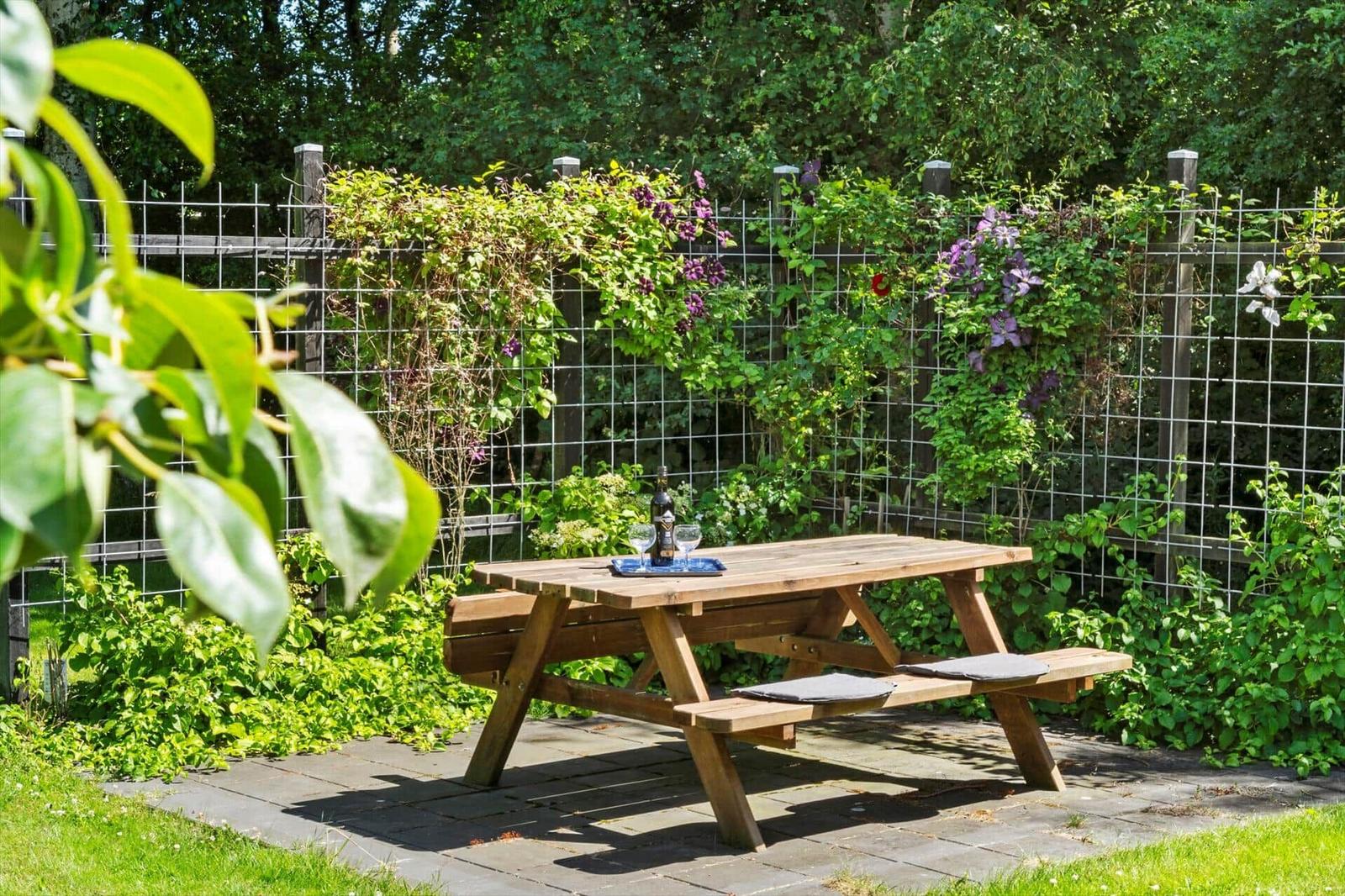 Holzpicknicktisch mit Flasche und Gläsern auf Steinplatten. Hintergrund: Ranken mit lila Blüten an Zaun.