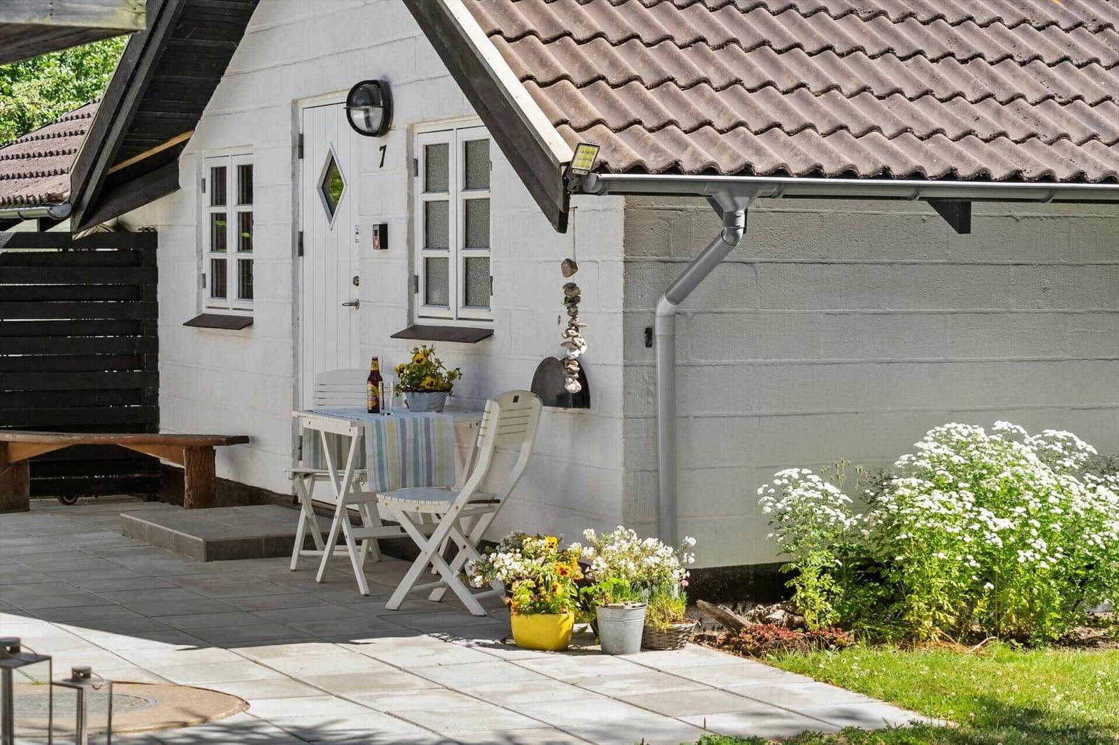 Weiße Hütte mit Terrasse, Tisch, Stühlen und Blumenbeeten. Holztor und Ziegeldach sichtbar.