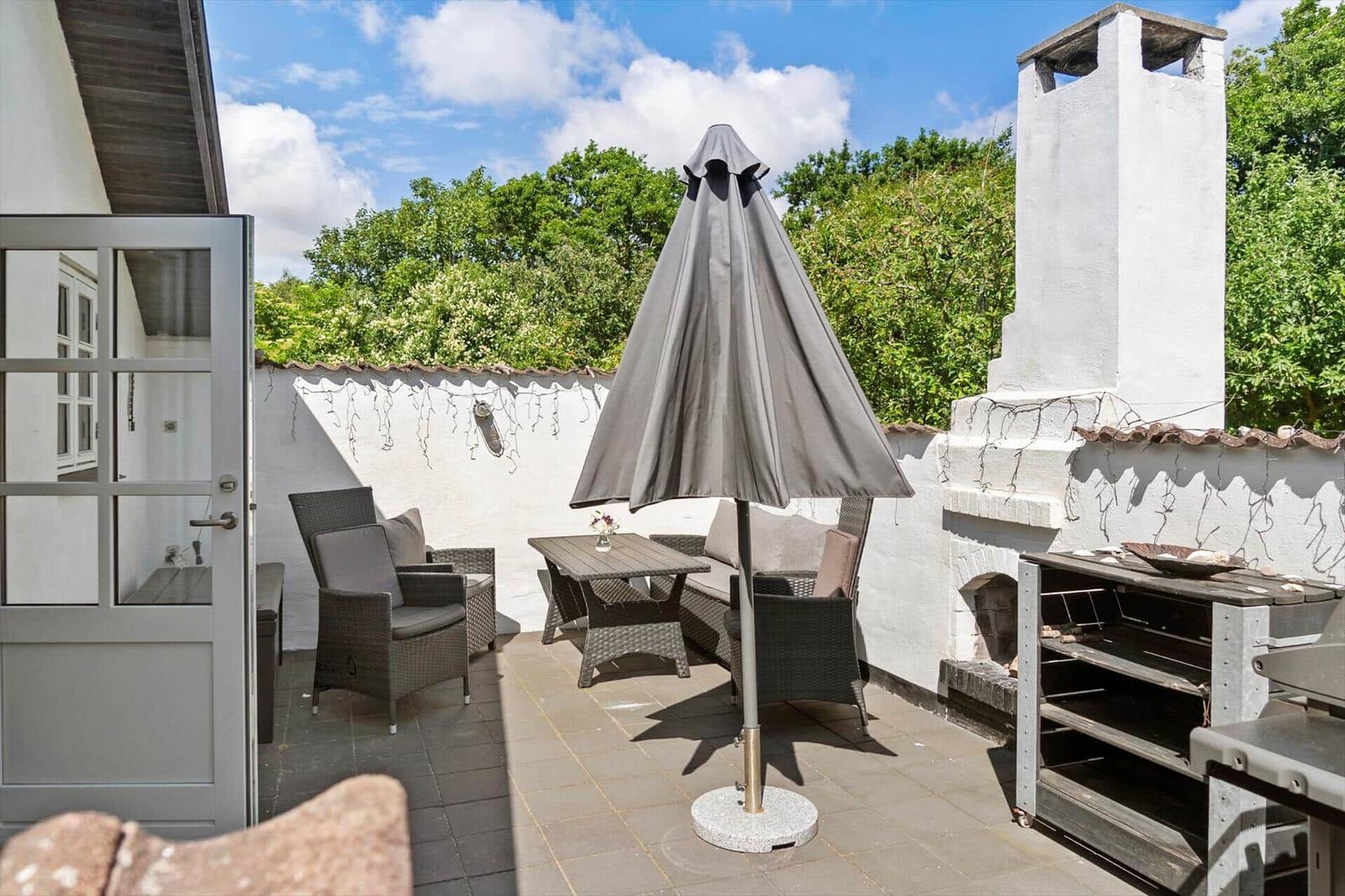 Terrasse mit Sitzgruppe, Sonnenschirm und Grillbereich. Weiße Mauer und grüne Bäume im Hintergrund.