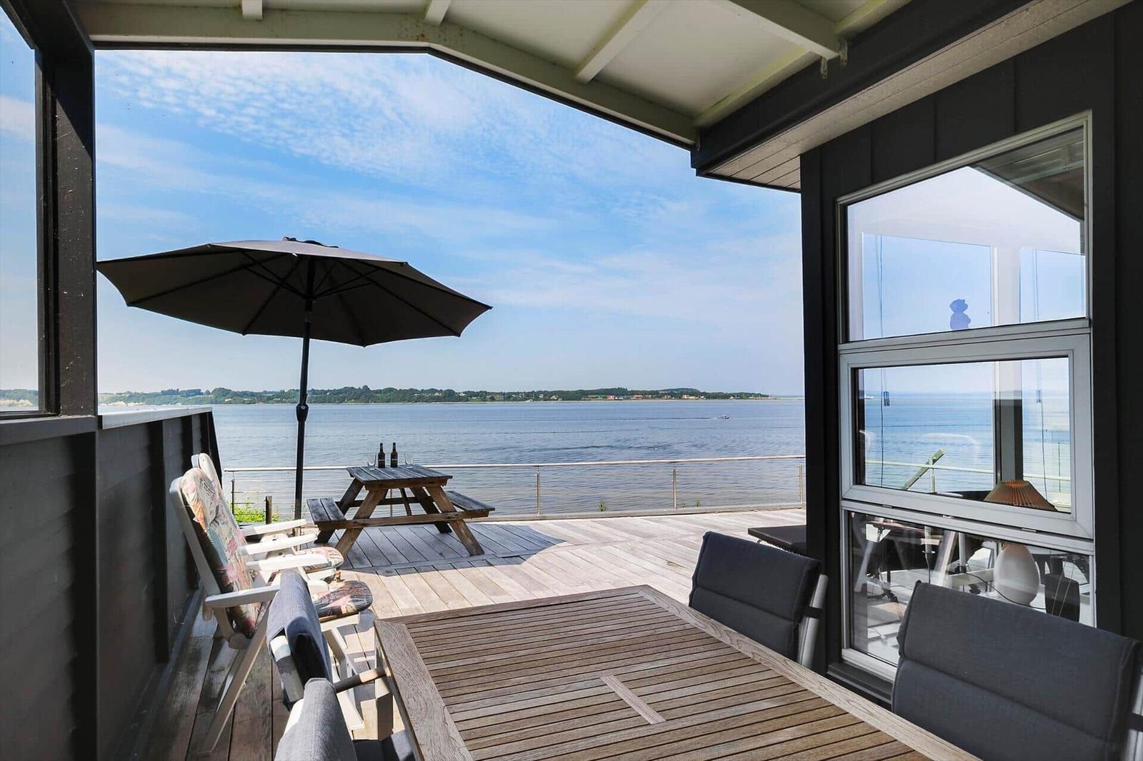Terrasse mit Tisch, Stühlen und Sonnenschirm. Blick auf ruhiges Wasser und gegenüberliegende Ufer.