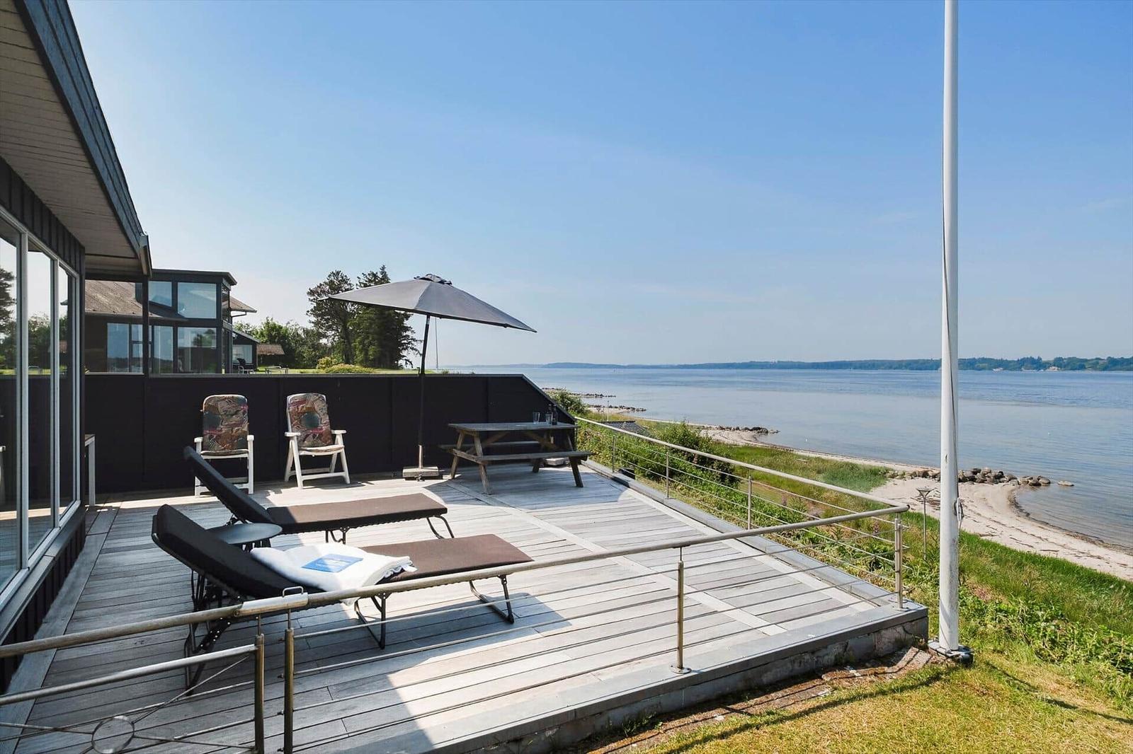 Terrasse mit Liegestühlen, Tisch und Sonnenschirm am Wasser.