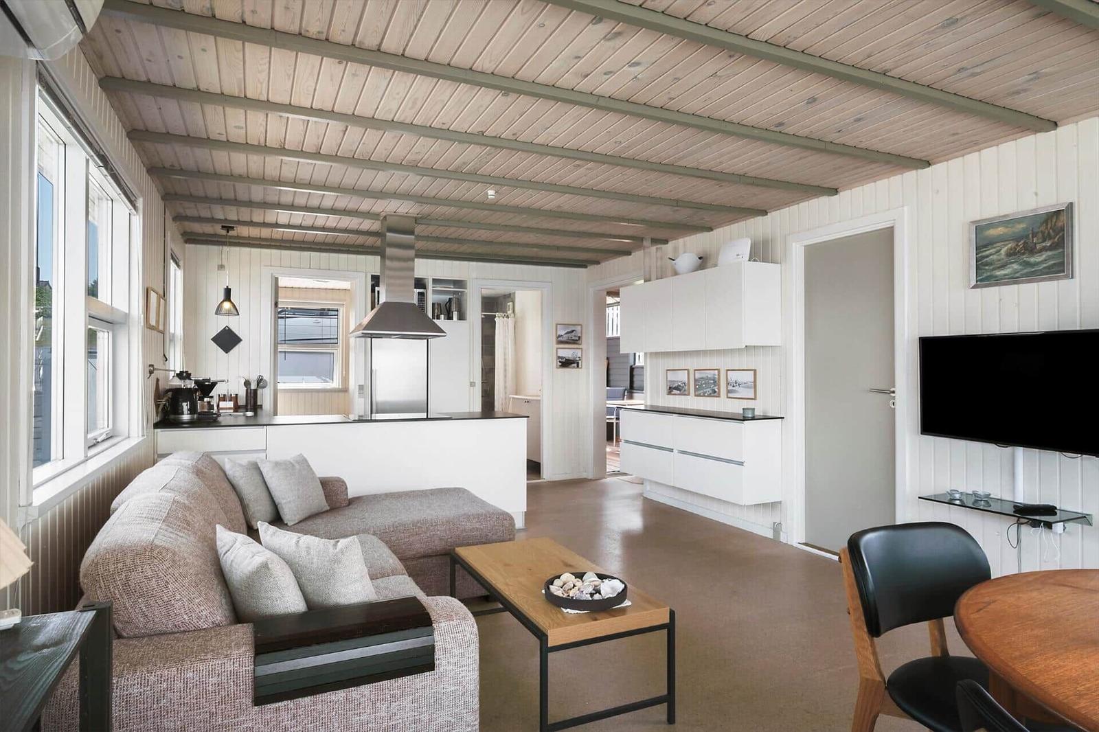 Wohn- und Küchenbereich mit Sofa, Esstisch und Fernseher. Holzdecke und weiße Wände.