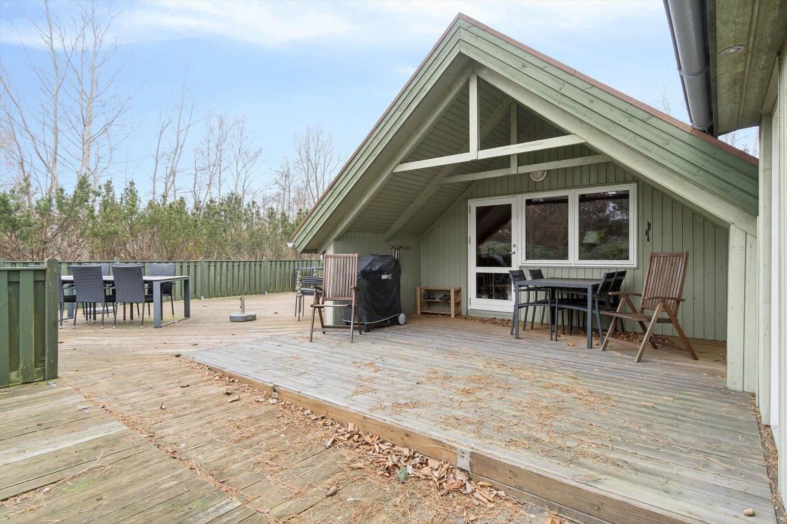 Holzterrasse mit Grill, Tischen und Stühlen vor einem Haus mit Dachüberstand.