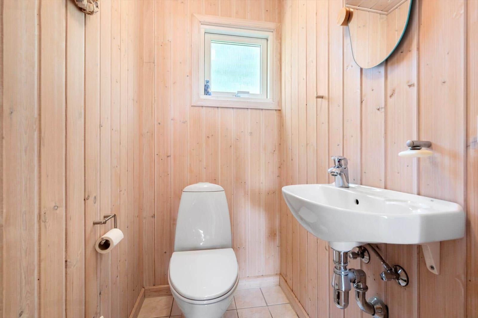 Badezimmer mit Toilette, Waschbecken und kleinem Fenster. Holzwandverkleidung und Fliesenboden.