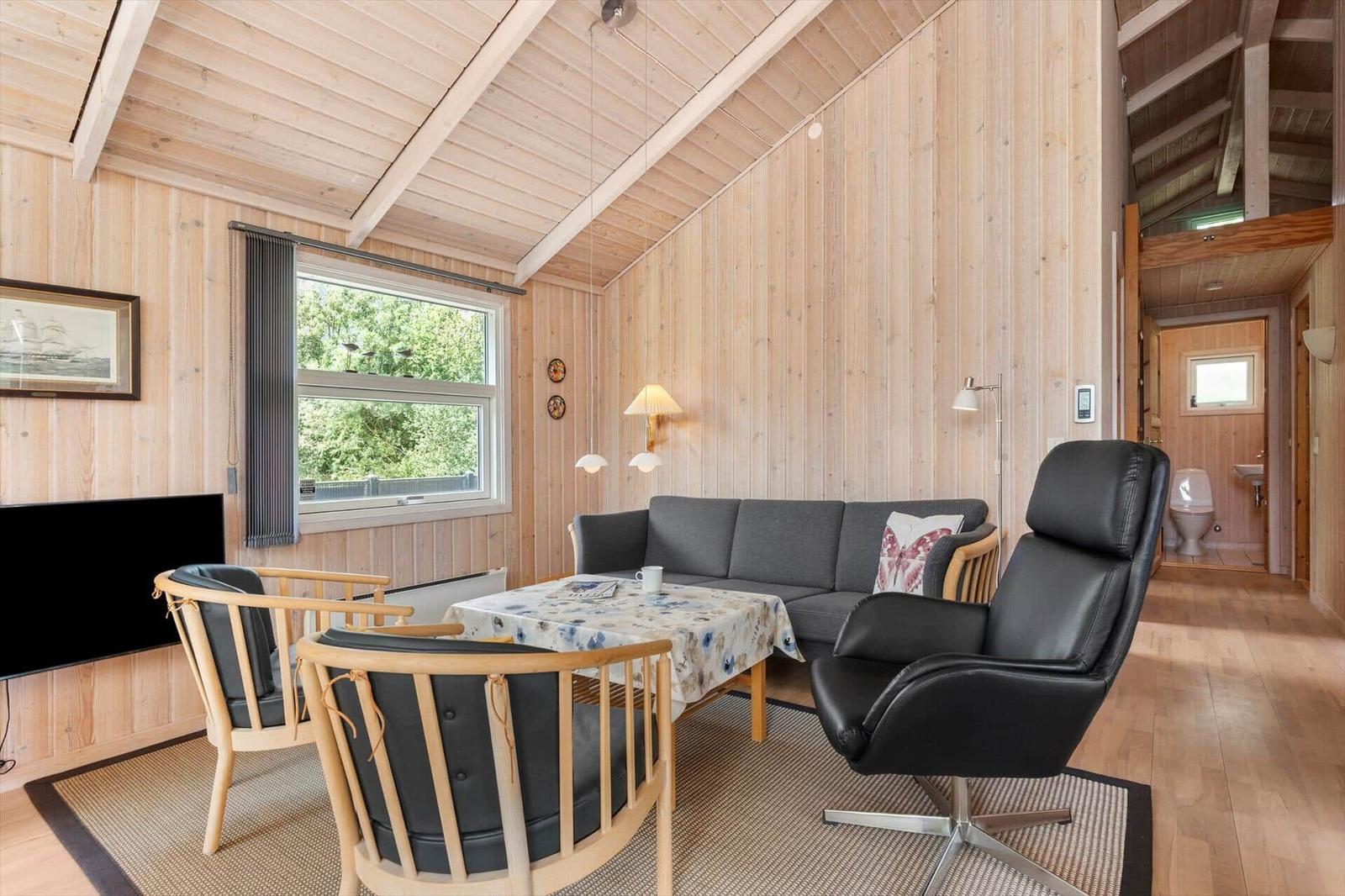 Wohnzimmer mit Sofa, Sessel und Tisch. Holzwände und -decke. Fenster mit Blick auf Grün.
