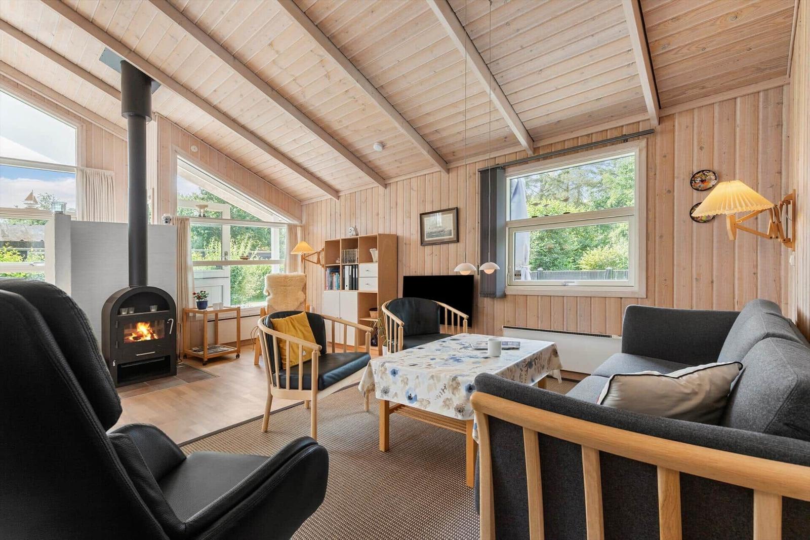 Wohnzimmer mit Holzdecke, Kamin, Sofa und Sitzgelegenheiten. Große Fenster mit Blick auf Garten.