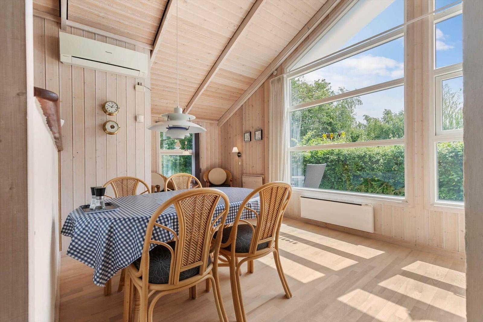 Esszimmer mit Holzwand, Tisch mit Tischdecke und Stühlen, großes Fenster mit Blick auf Grün.