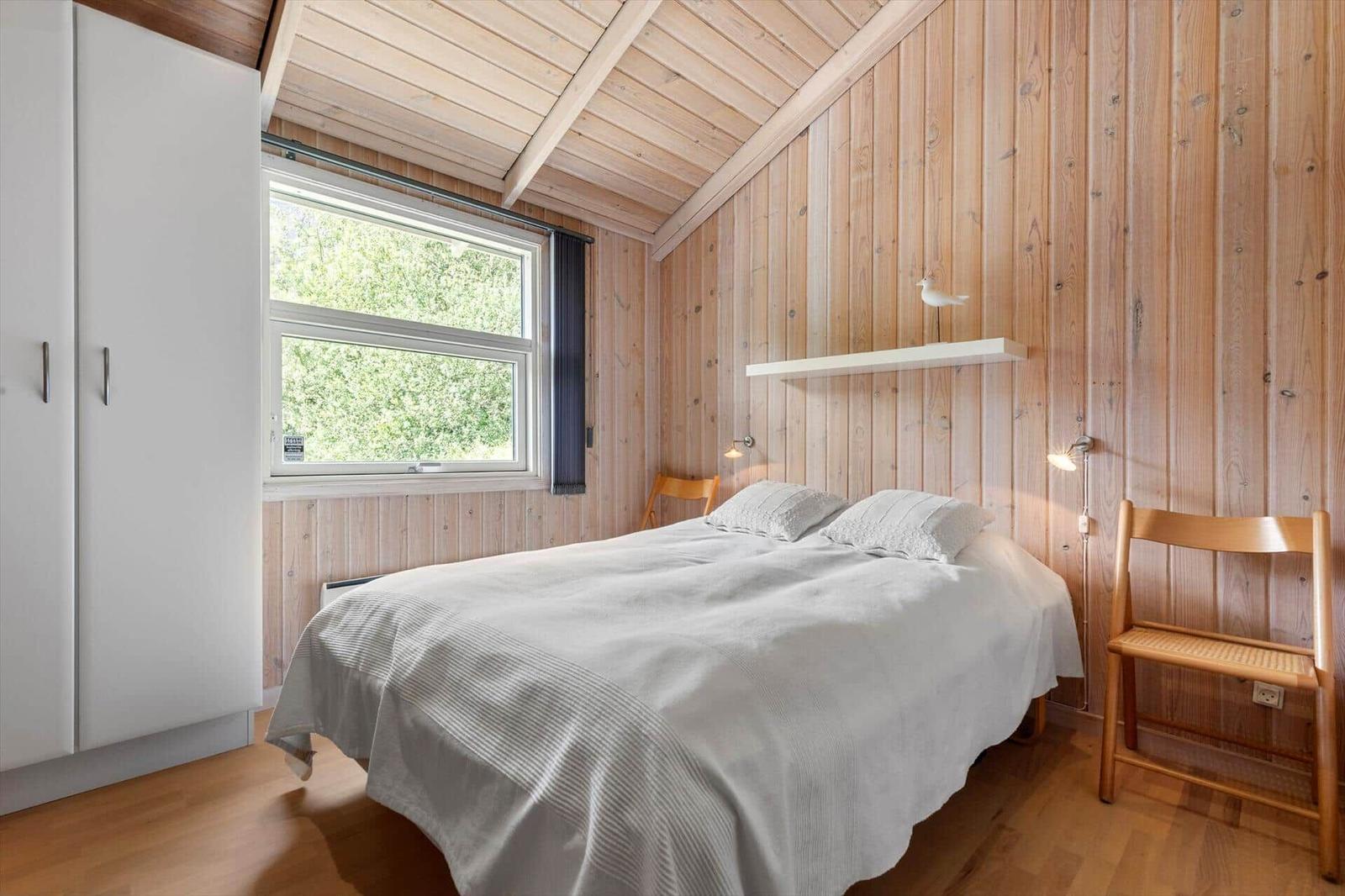 Schlafzimmer mit Bett, Schrank und Stuhl. Holzwände und Fenster mit Blick auf Grün.
