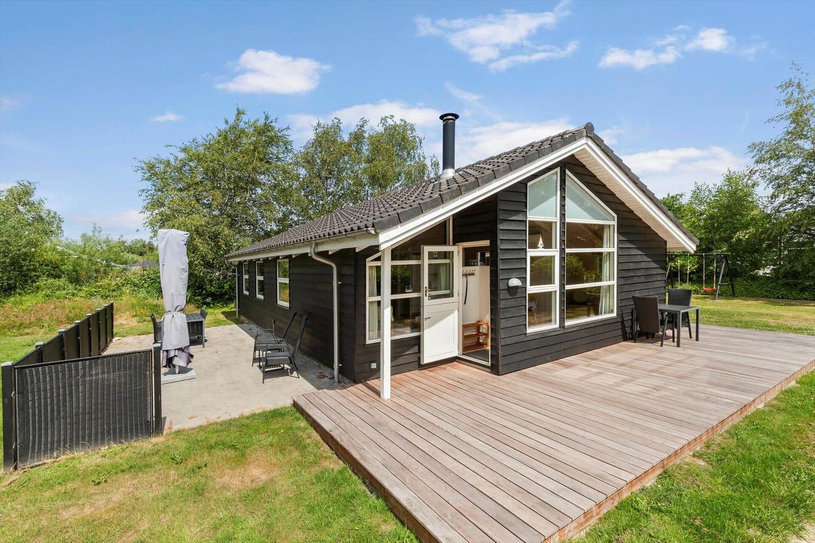 Schwarzes Holzhaus mit Terrasse und Garten. Große Fenster, Außentisch und Schaukel.