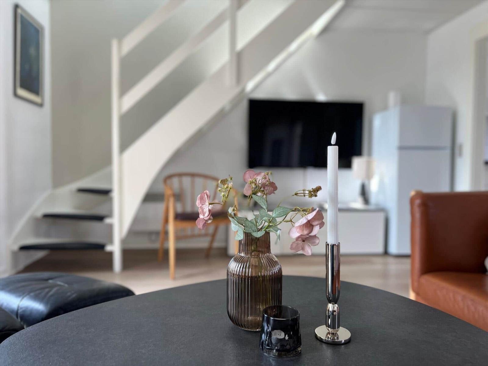 Runde Tisch mit Blumen, Kerze und Glas. Im Hintergrund Stuhl, Fernseher und Kühlschrank.