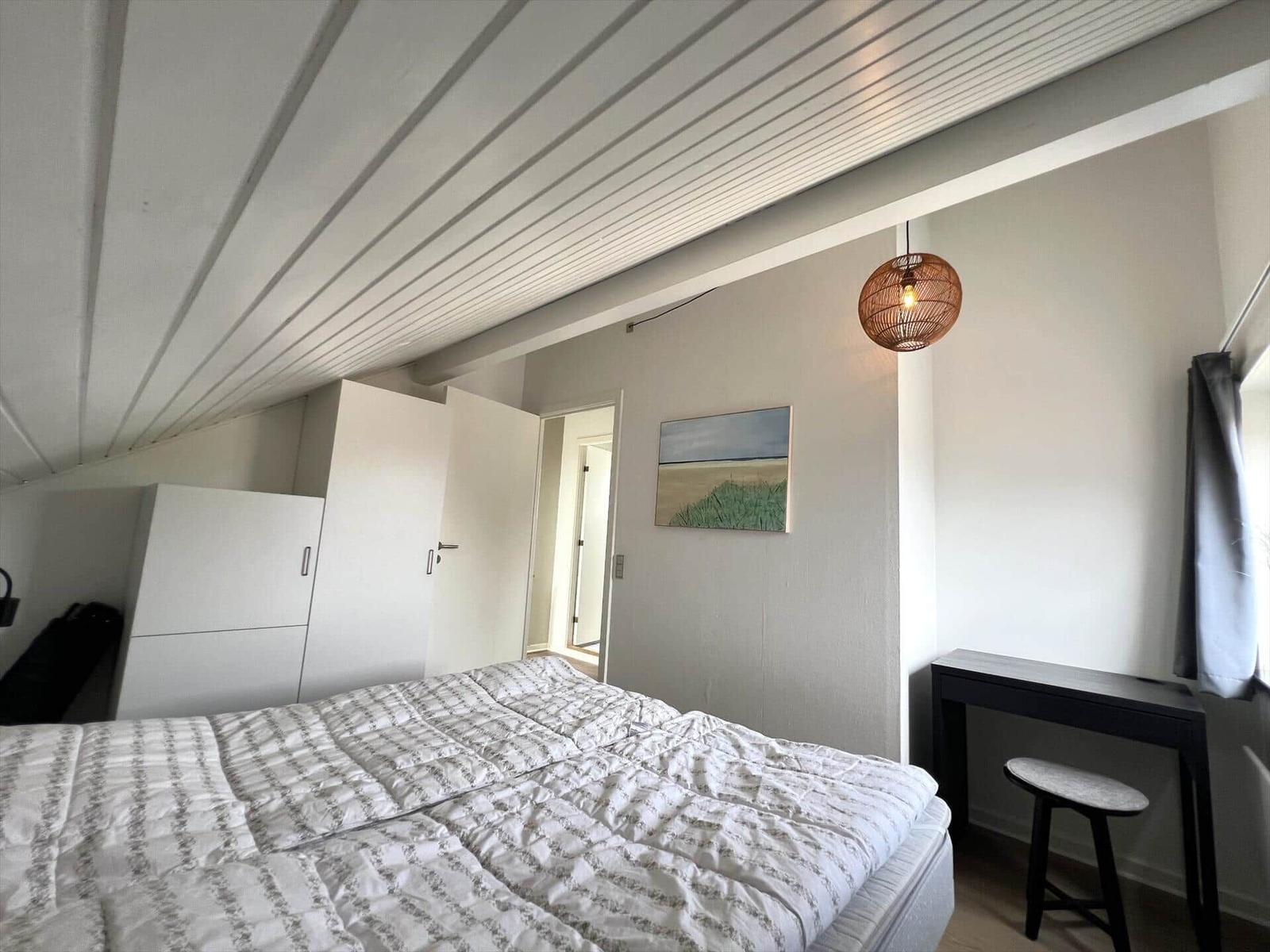 Schlafzimmer mit Bett, Kleiderschrank, Schreibtisch und Pendelleuchte unter schrägem Dach.