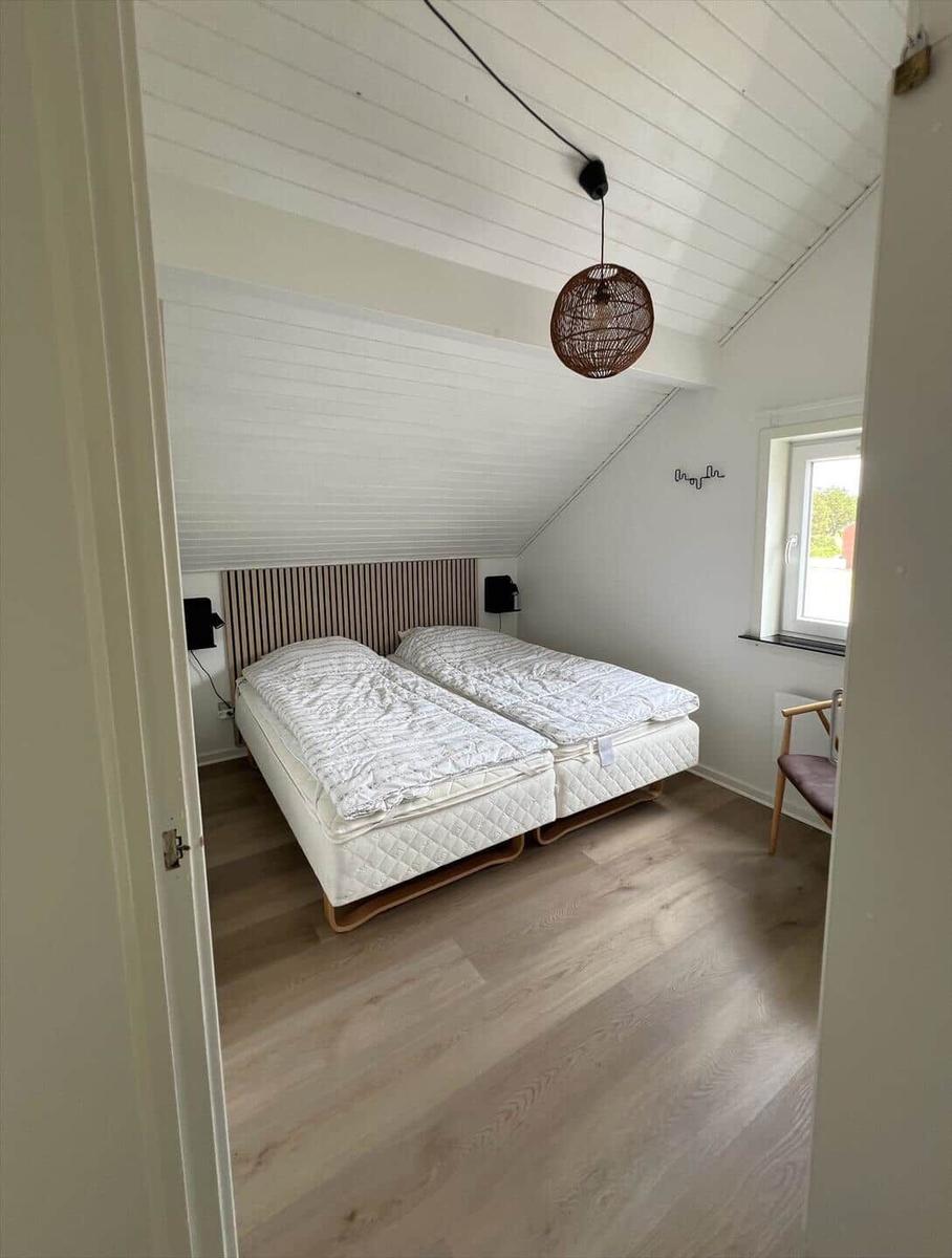 Schlafzimmer mit Doppelbett, Holzboden und schrägem Dach. Fenster und Hängelampe sichtbar.
