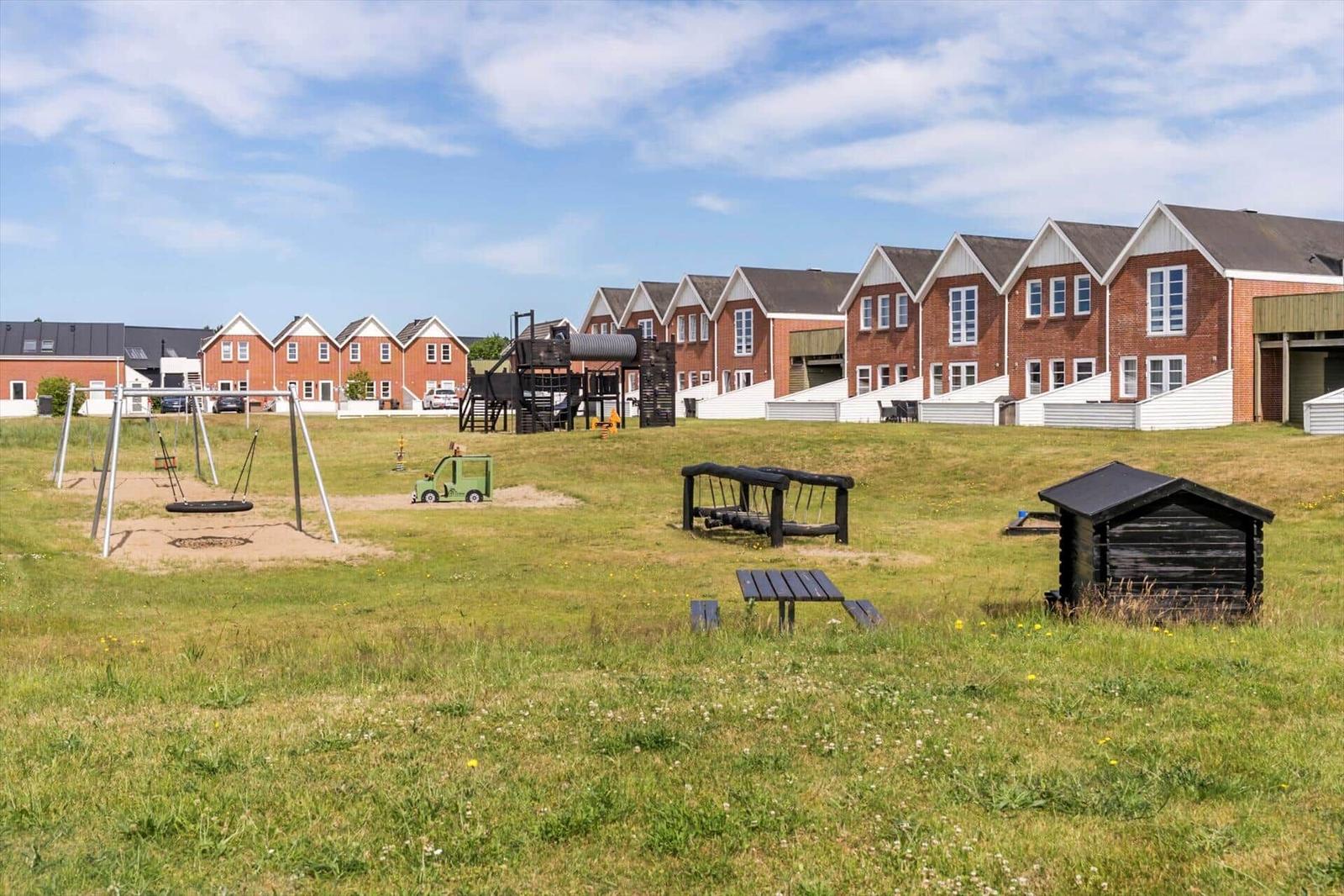 Grüner Spielplatz mit Schaukel, Rutsche und Holzhütte vor Reihenhäusern.