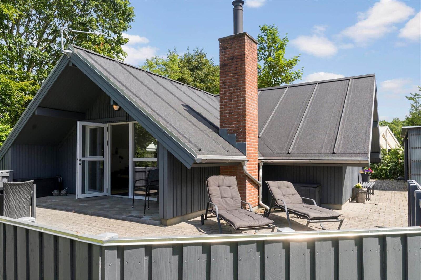 Haus mit Terrasse, Liegestühlen und Schornstein. Holzverkleidung und graue Dächer.