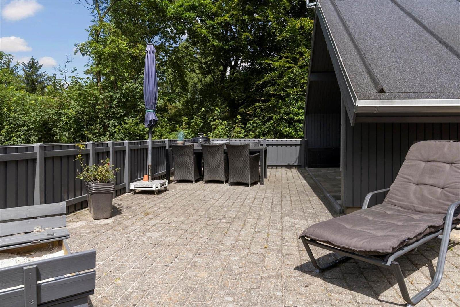 Terrasse mit Liegestuhl, Tisch, Stühlen und Sonnenschirm. Hintergrund: grüne Bäume und Holzzaun.