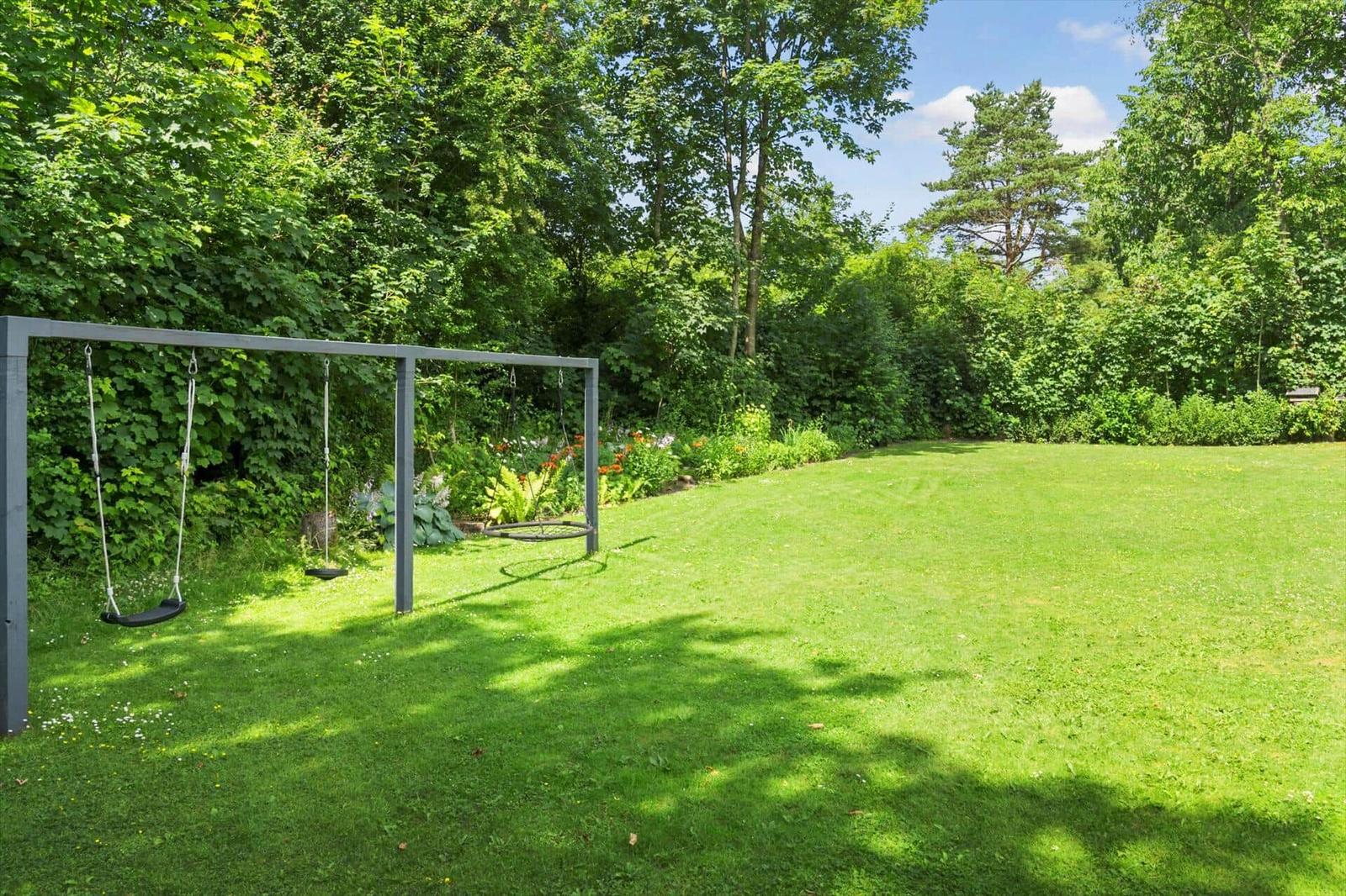 Großer Garten mit Schaukelanlage und grünem Rasenbereich. Umgeben von Bäumen und Sträuchern.