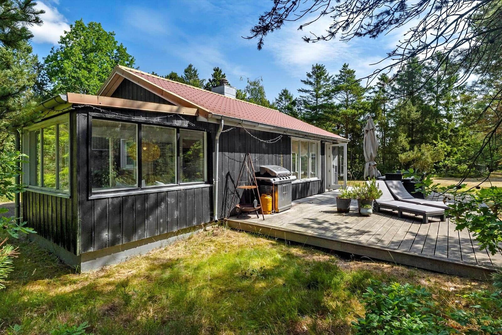 Ein Haus mit Holzveranda, Grill und Liegen im Grünen.