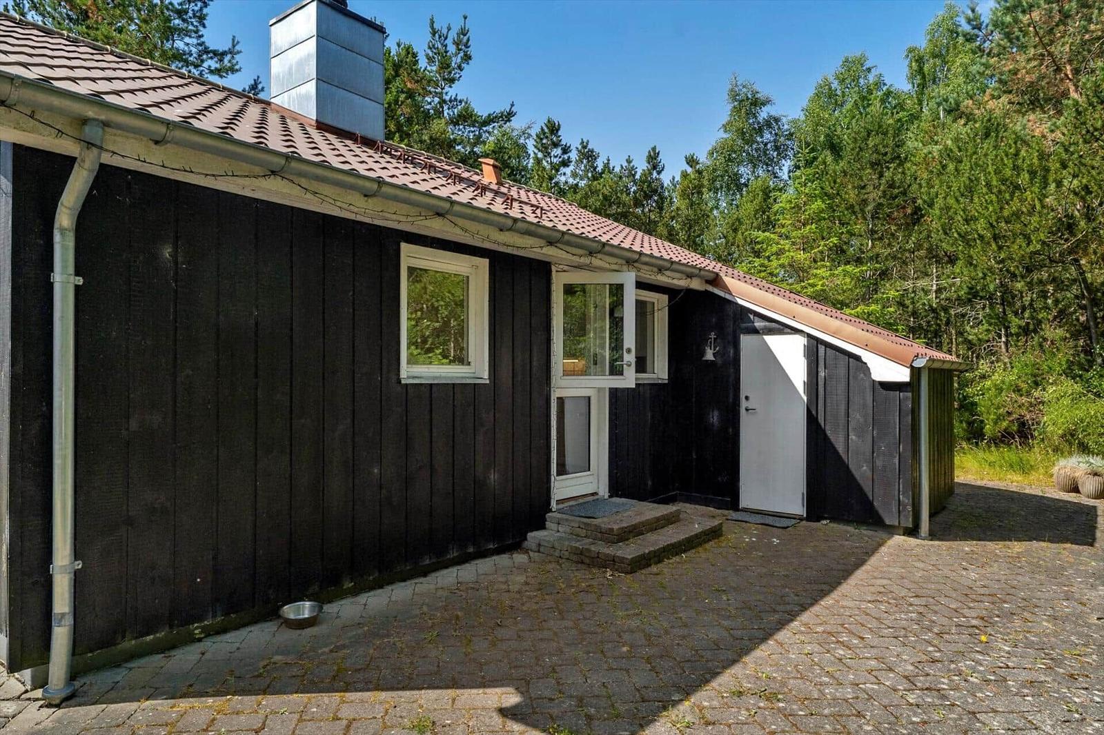 Schwarze Holzhütte mit rotem Dach, weißer Tür und Fenster. Umgeben von Wald und gepflastertem Hof.