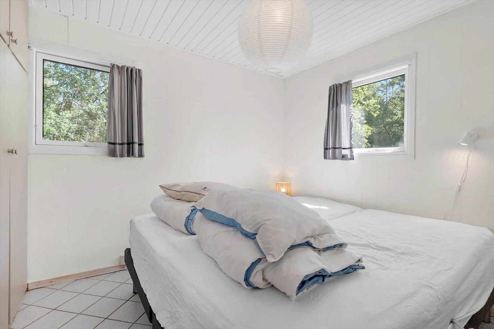 Schlafzimmer mit Bett, zwei Fenstern und einer Deckenleuchte.