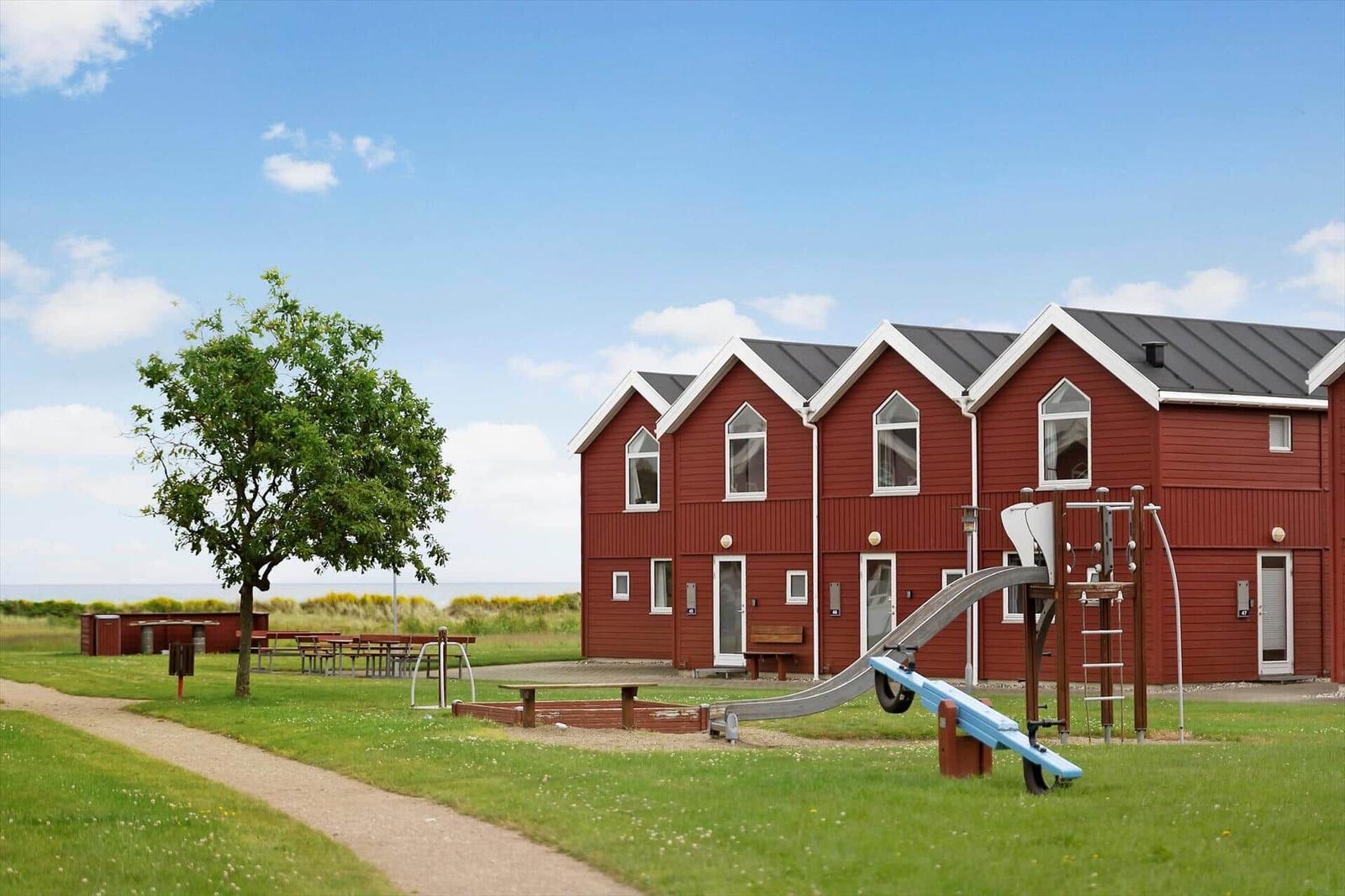 Rote Holzhäuser mit Spielplatz und Grünfläche. Strand im Hintergrund.