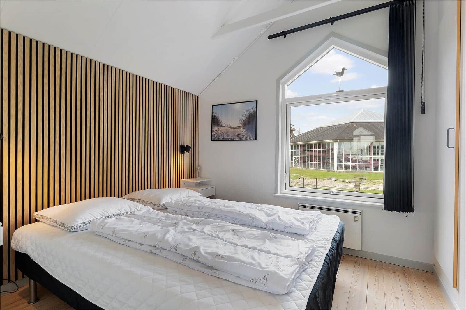 Schlafzimmer mit Holzwand, großem Fenster und Bett mit weißer Bettwäsche.