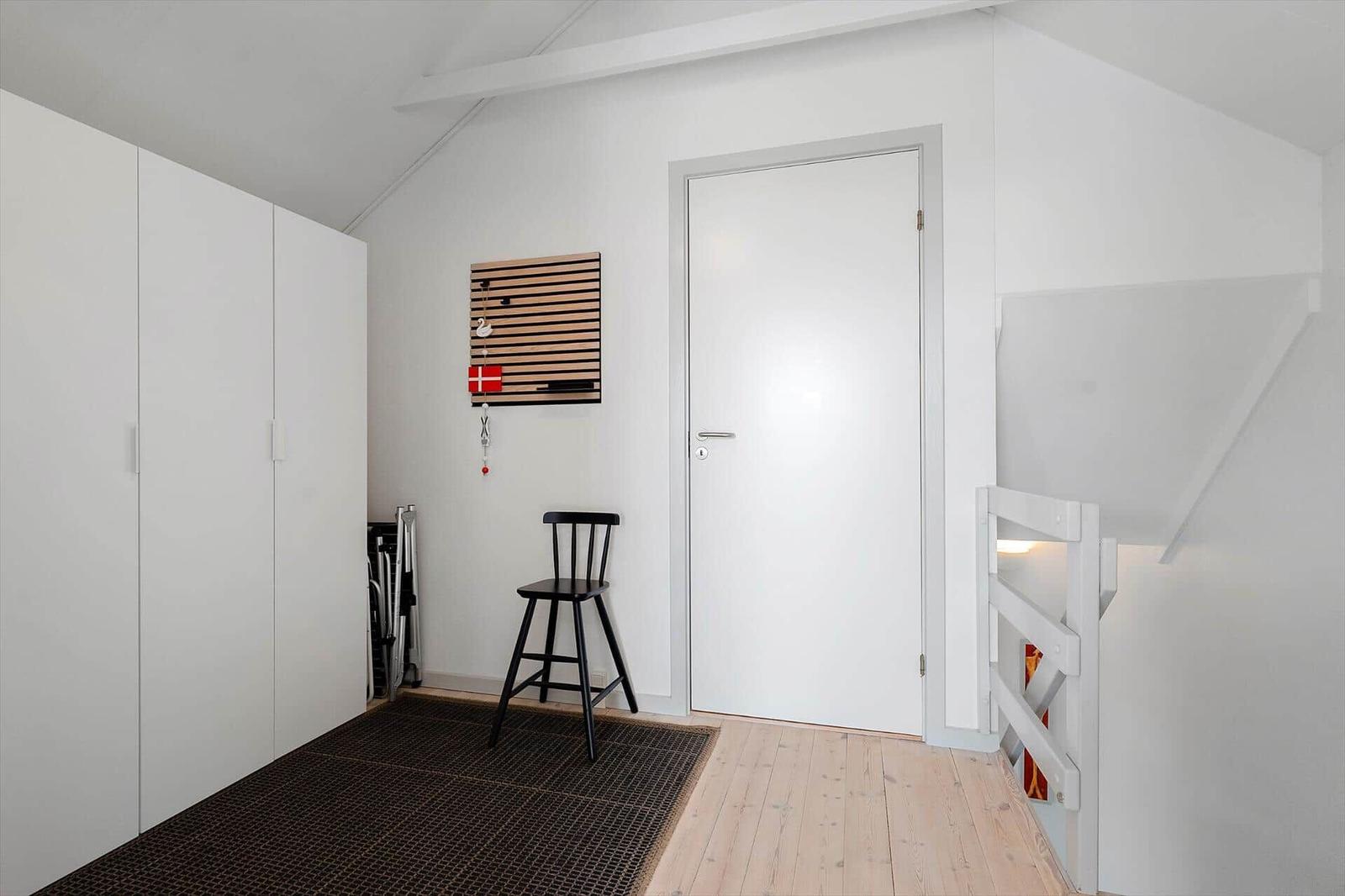 Raum mit weißen Wänden, Holzboden, Schrank und Stuhl. Tür und Treppe sind ebenfalls weiß.