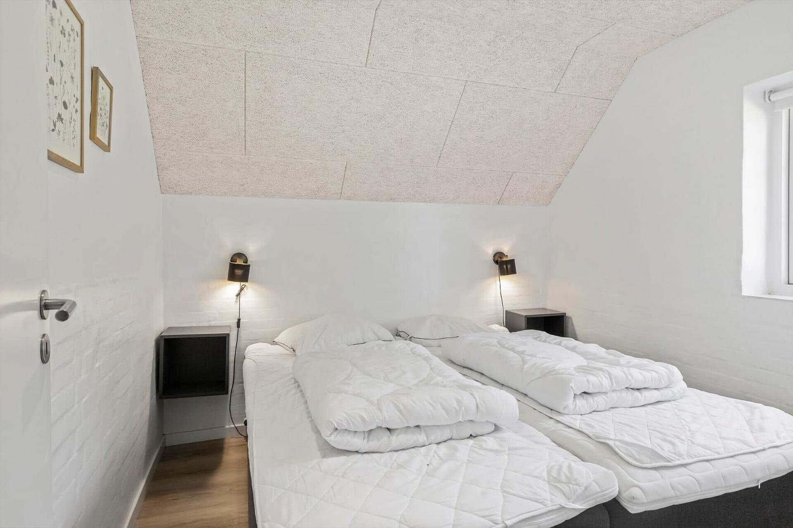 Zwei Betten mit weißen Bettwäsche unter schrägem Dach mit Wandlampen und Fenster.