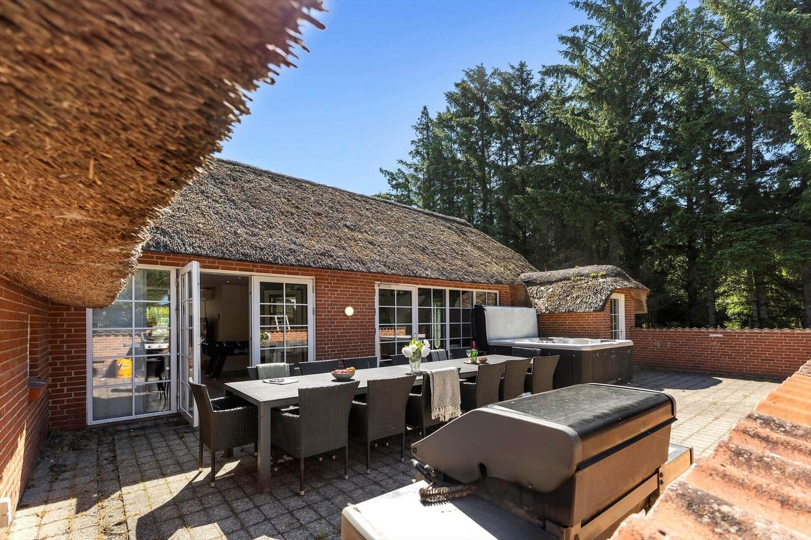 Terrasse mit Esstisch, Grill und Whirlpool. Roter Backsteinbau mit Strohdach und Bäumen im Hintergrund.