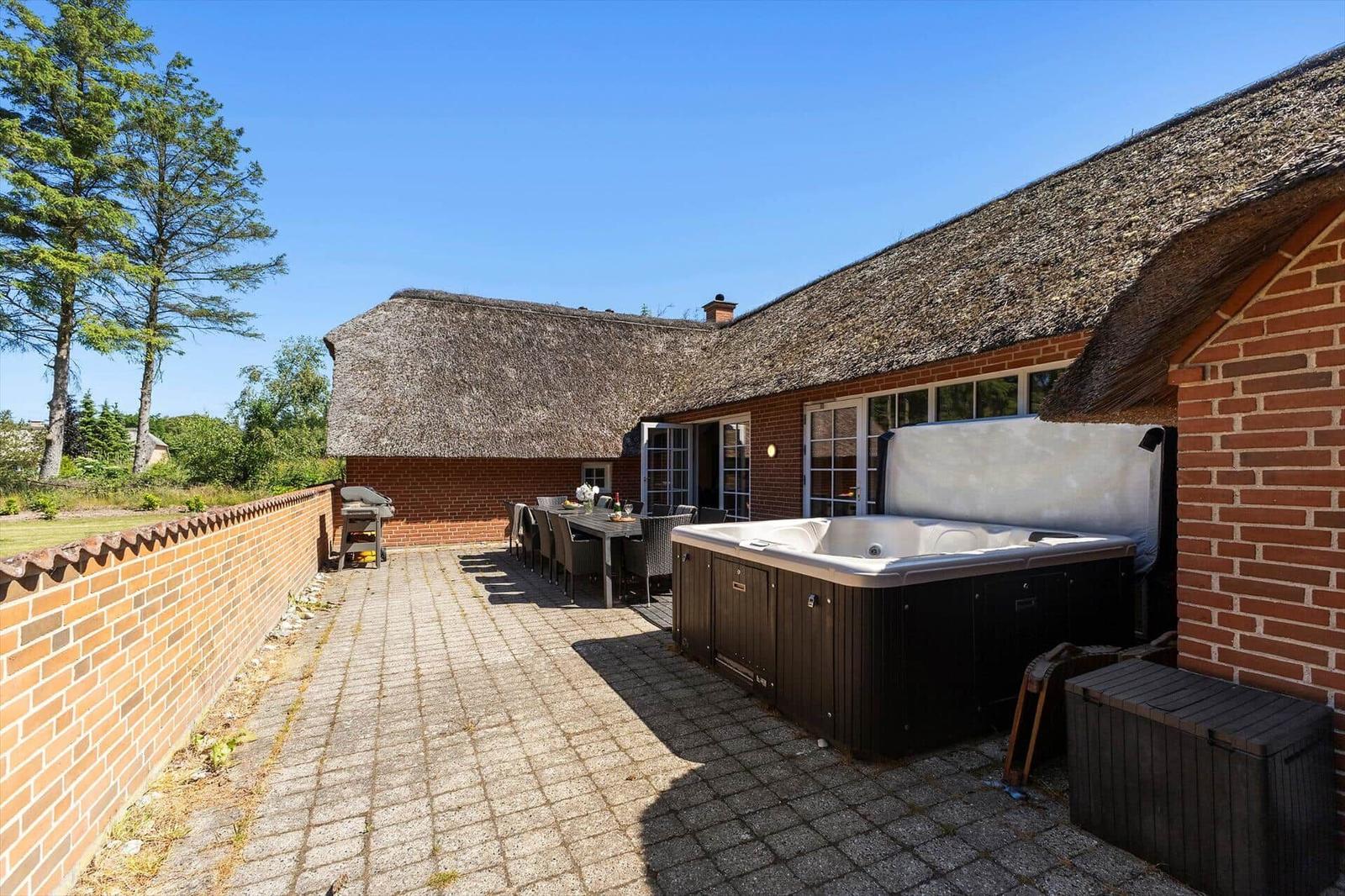 Terrasse mit Whirlpool, Grill und Tisch. Holzhaus mit Strohdach und Ziegelmauer.