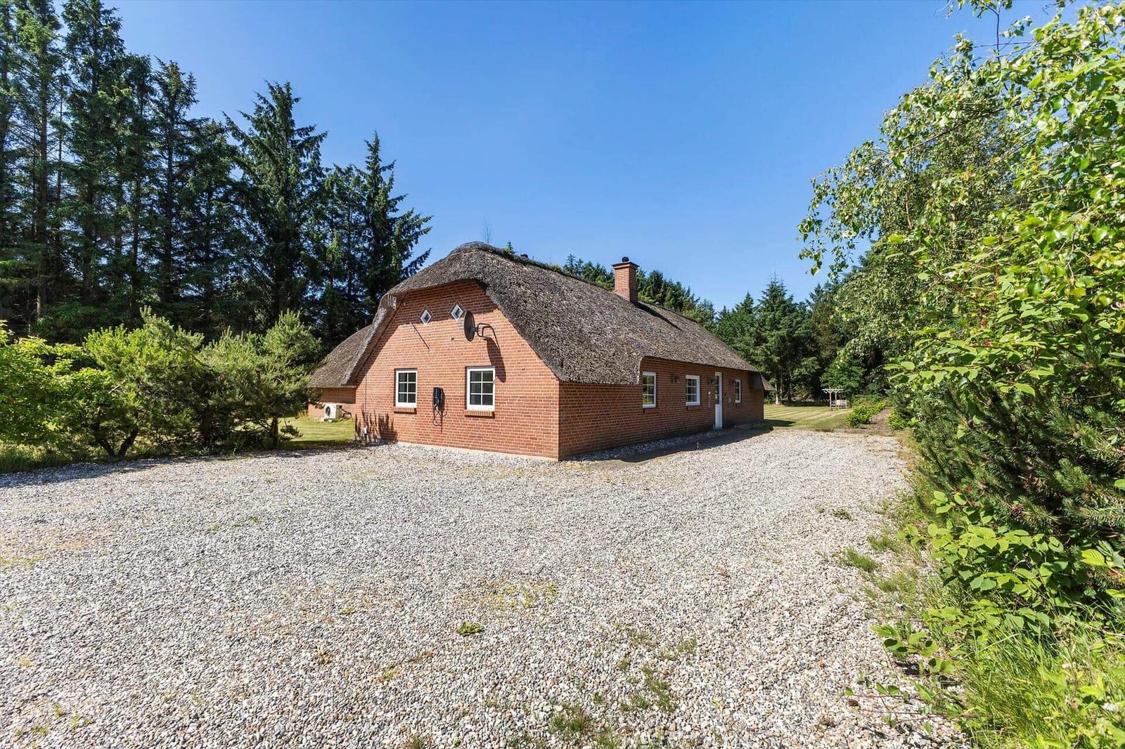 Ein rotes Backsteinhaus mit Strohdach steht auf einem Kiesplatz umgeben von Bäumen.