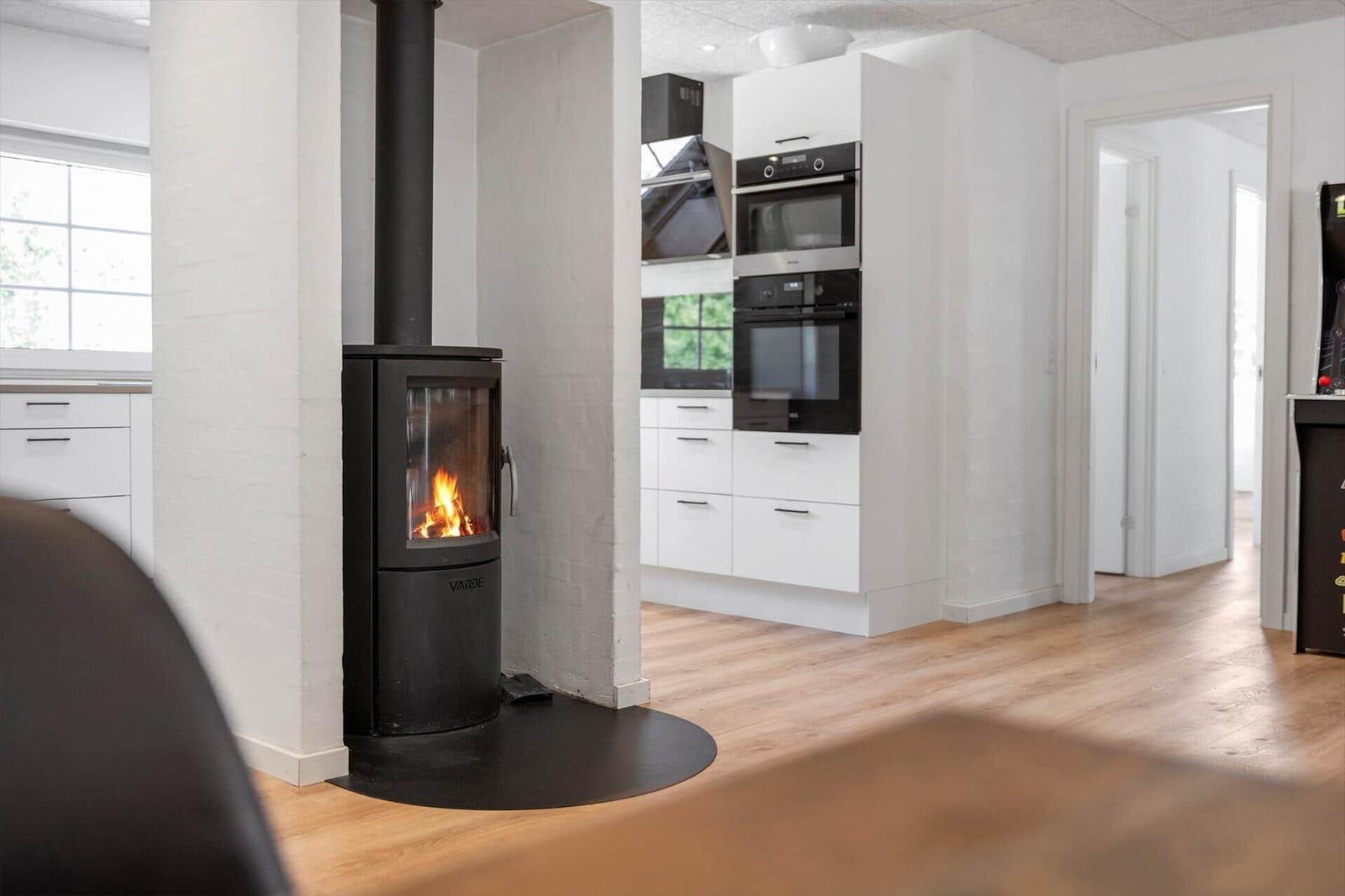 Küche mit weißen Schränken und integrierten Geräten. Im Vordergrund steht ein Kaminofen mit brennendem Holz.