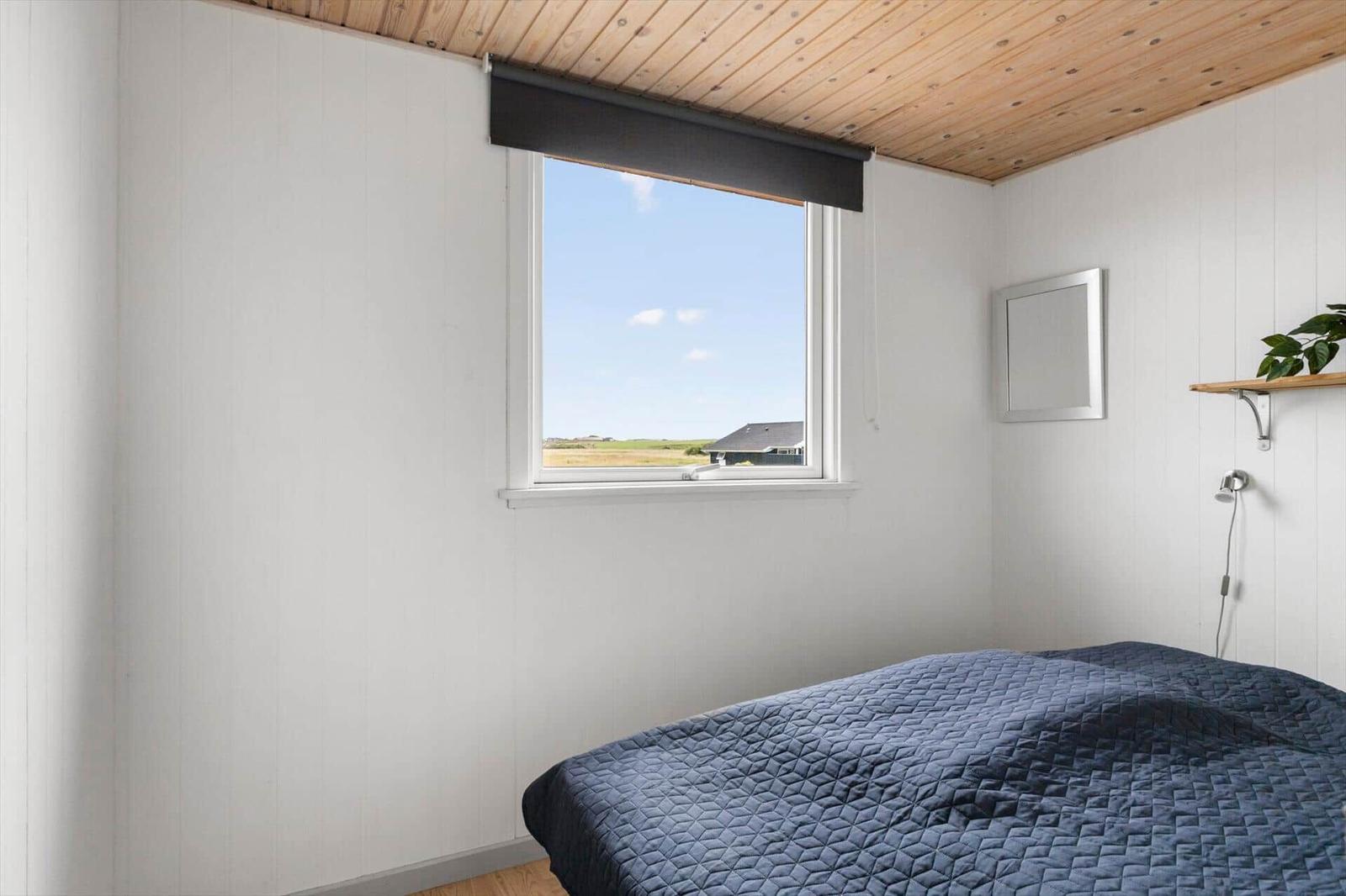 Schlafzimmer mit Bett, Fenster und Holzdecke. Draußen ist Feld und Himmel sichtbar.