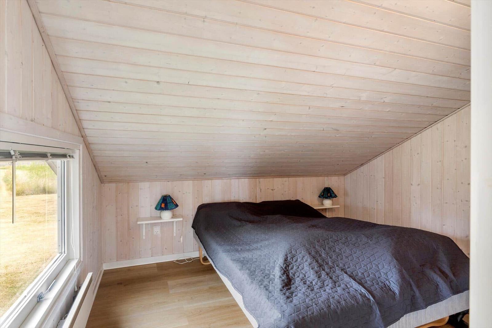 Schlafzimmer mit Holzwänden, Bett und Fenster. Zwei Nachttische mit Lampen.