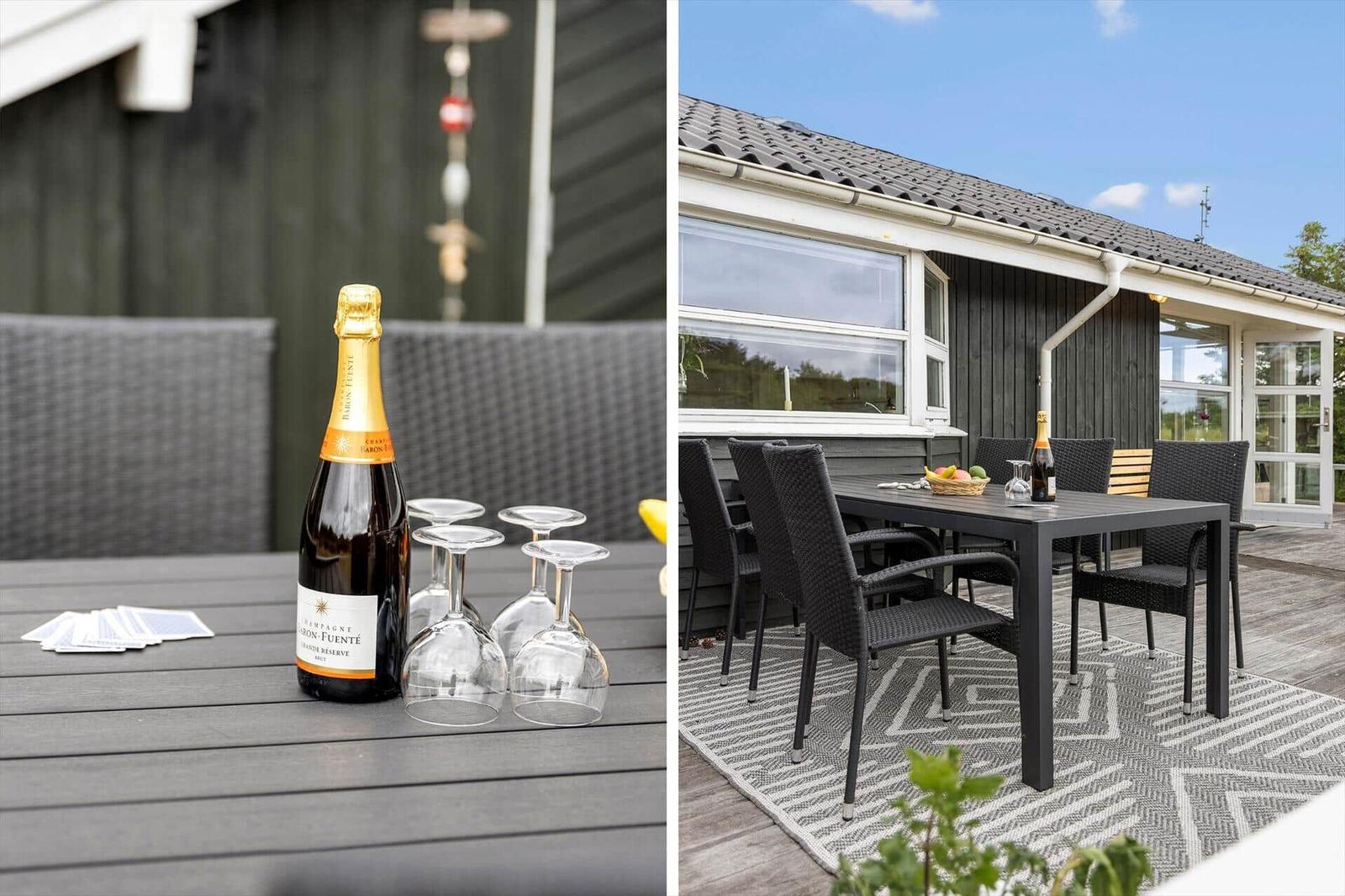 Außenbereich mit Tisch, Stühlen und Champagnerflasche. Haus mit dunkler Fassade und Terrasse.