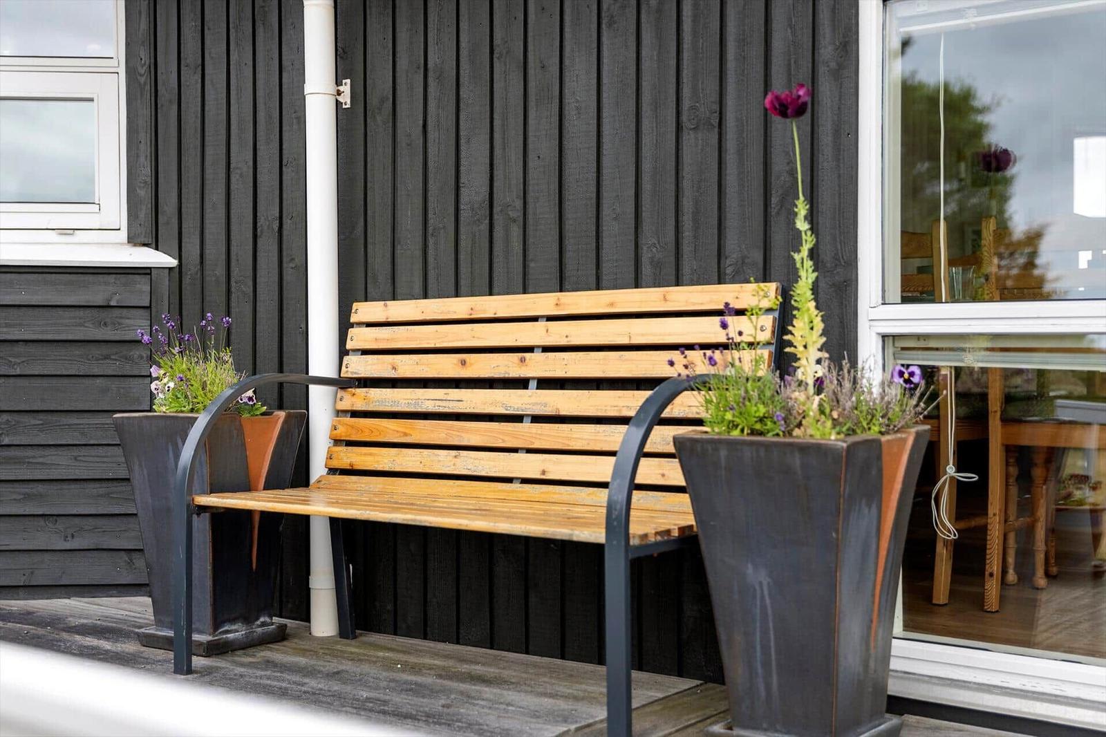 Holzbank mit zwei Pflanzenkästen auf einer Terrasse vor einem Haus mit dunklem Holzverkleidung.