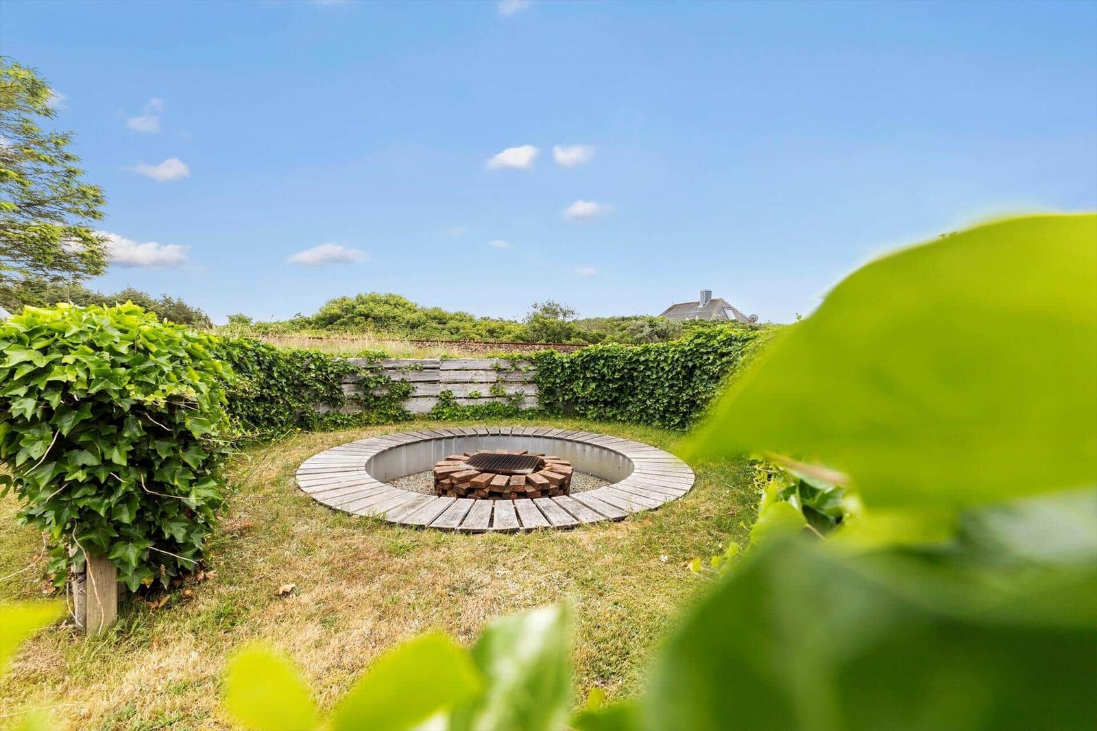 Runder Feuerplatz mit Holzbank im Garten, umgeben von Efeu und Gras.
