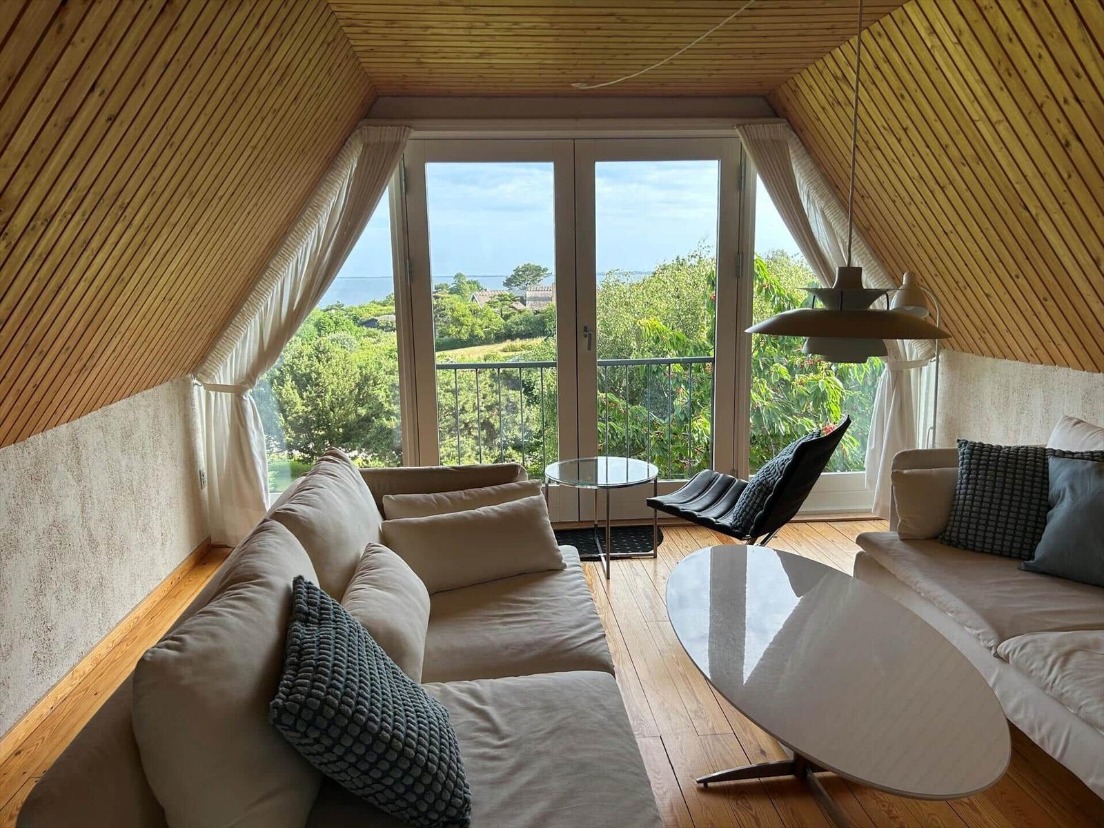 Living room with wooden ceiling, sofa, table, and balcony with view of greenery and sea.