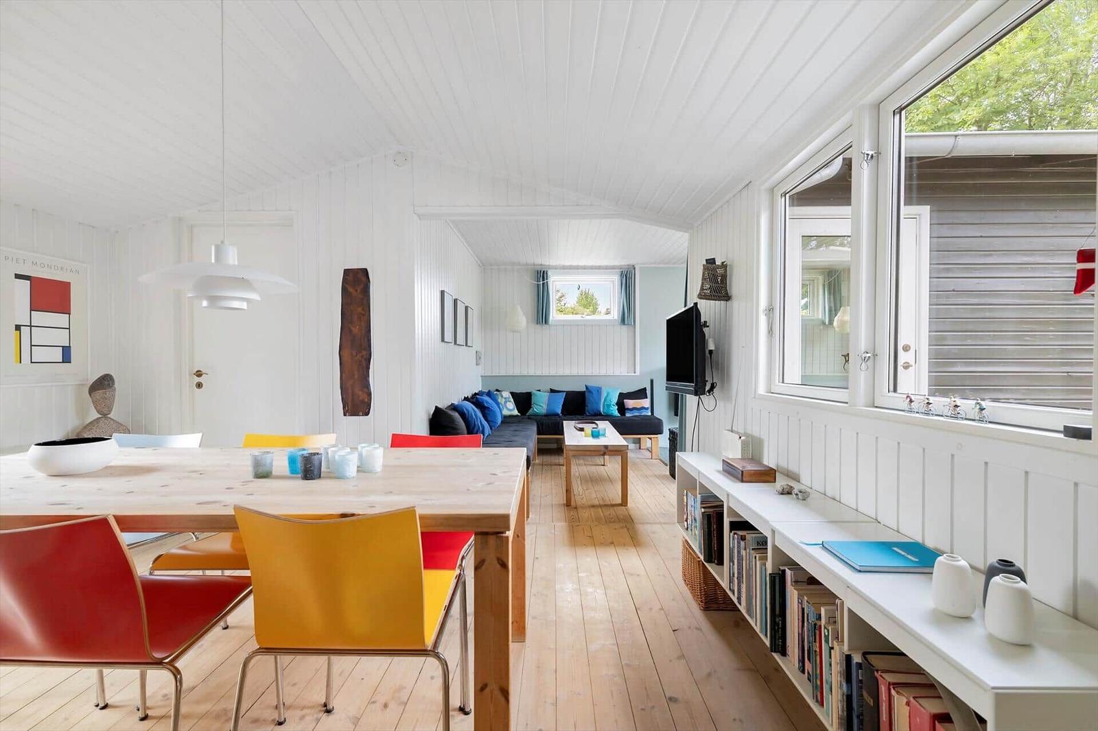 A room with dining and living areas. Wooden floor, white walls and windows. Colored chairs and sofa.