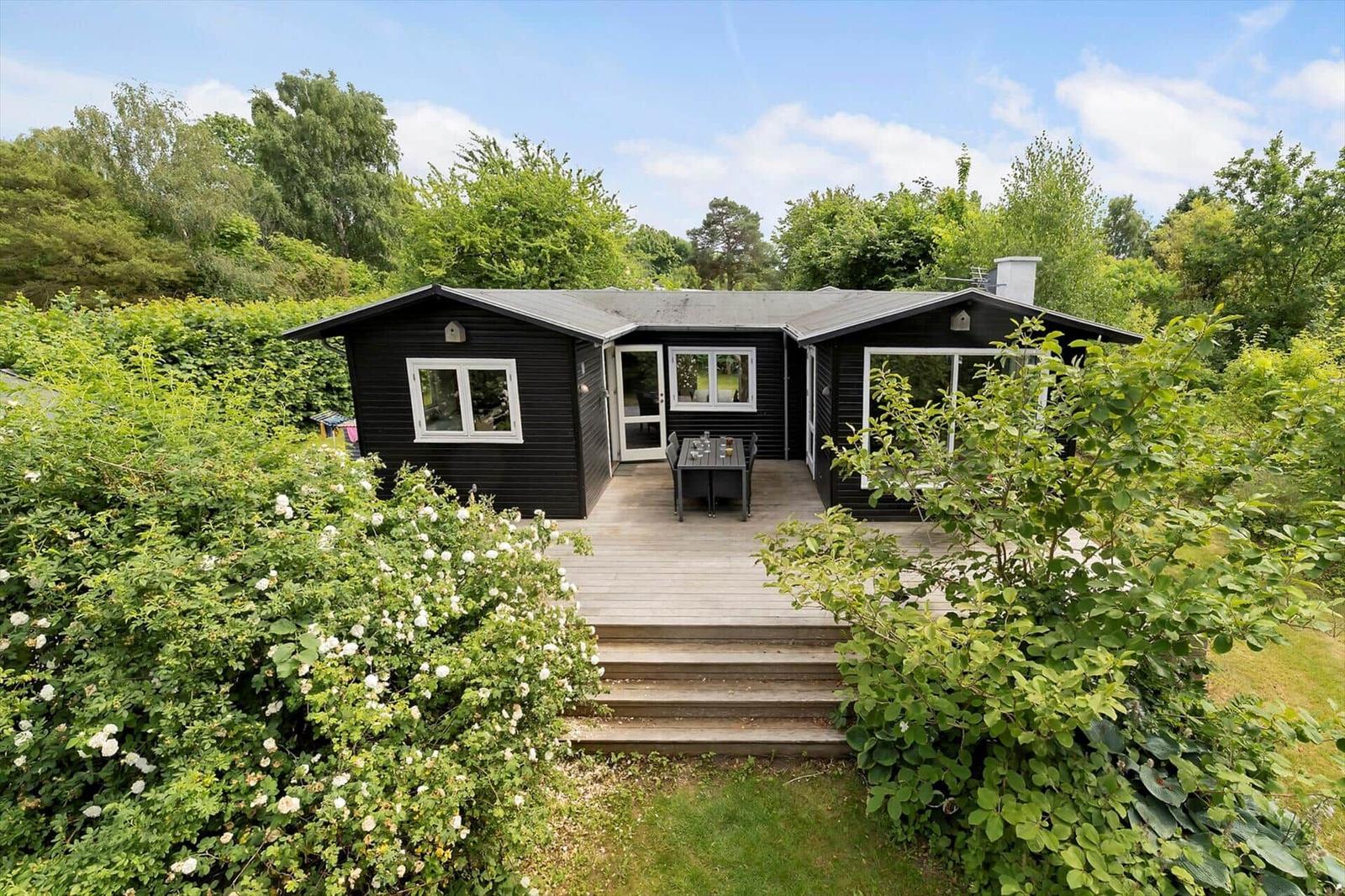 Black wooden house with terrace, garden, and trees in the background.