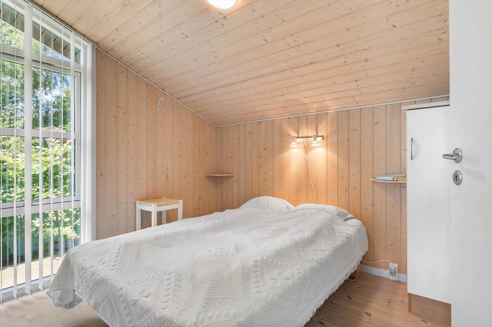 Schlafzimmer mit Holzwand und -decke, großes Fenster, Bett mit weißer Decke, Schrank und Wandlampe.