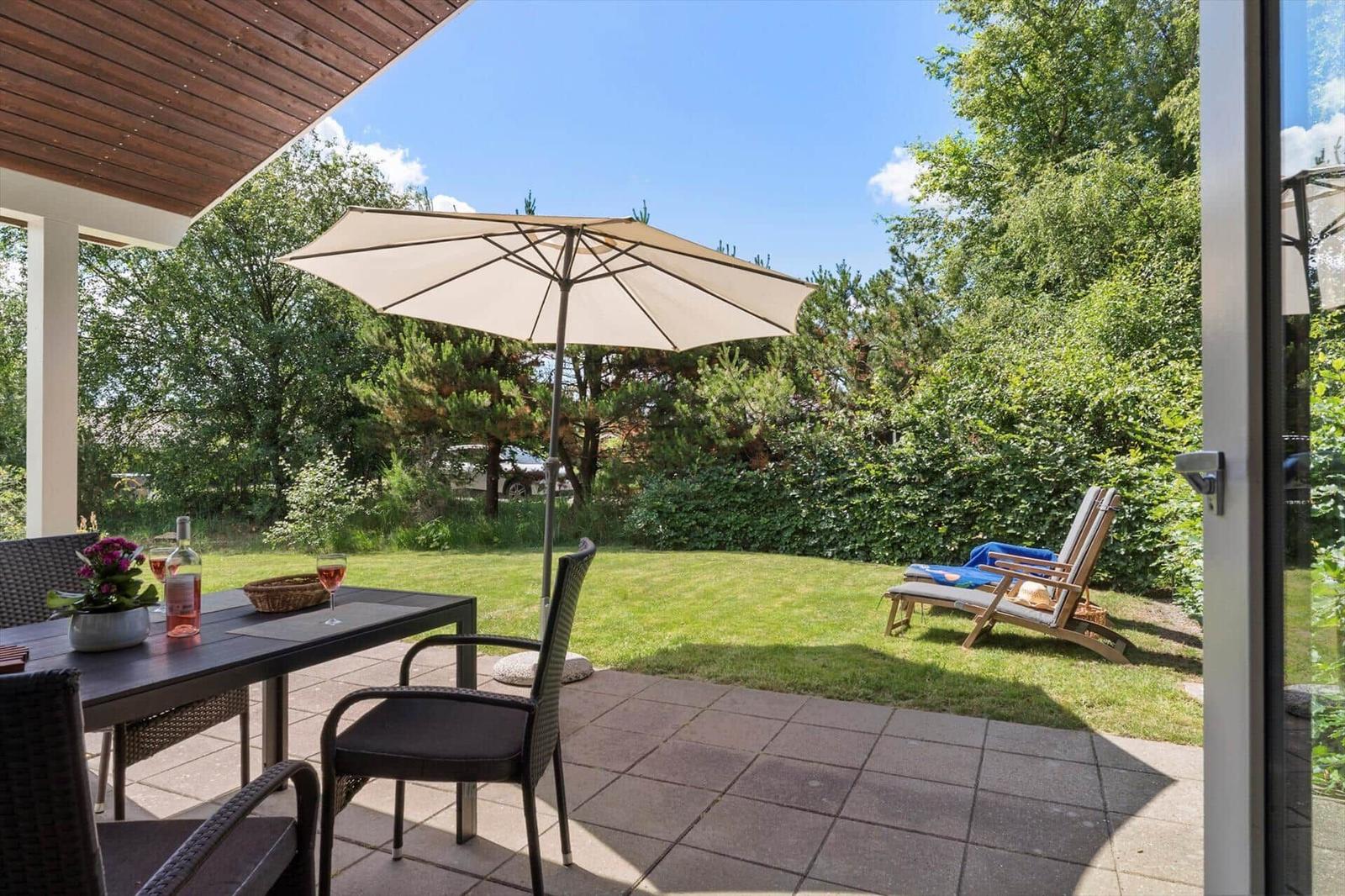 Terrasse mit Tisch, Stühlen und Sonnenschirm. Hintergrund: grüner Garten mit Liegestühlen.