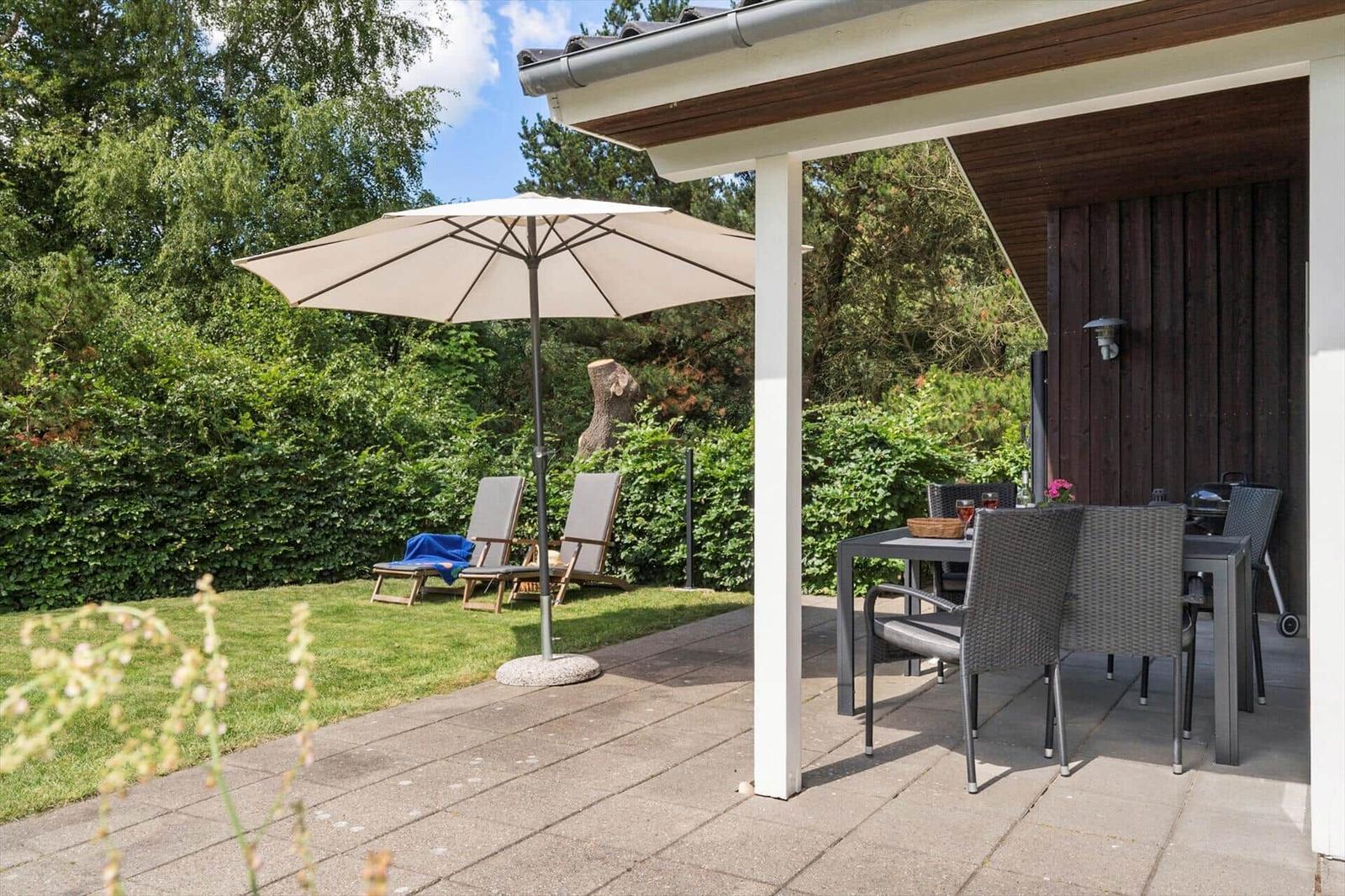 Terrasse mit Tisch, Stühlen und Sonnenschirm. Hintergrund: grüner Garten und Holzwand.
