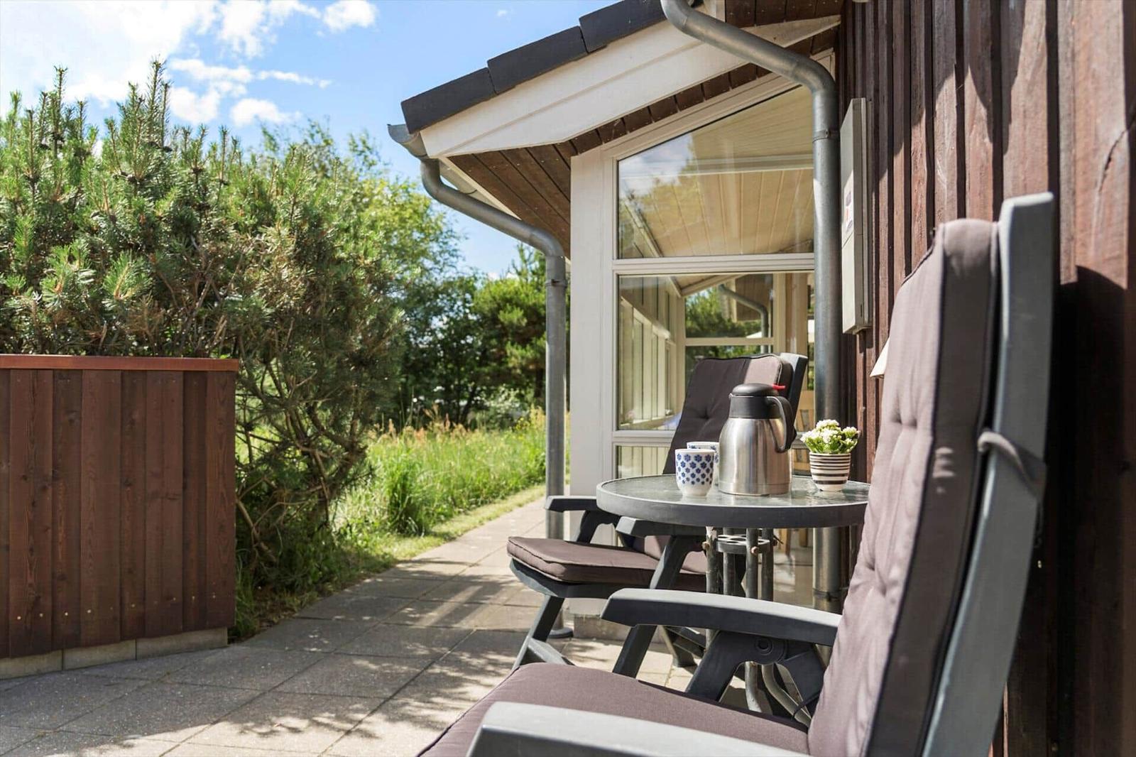 Terrasse mit Tisch, Stühlen und Thermoflasche. Hintergrund: Grünflächen und Holzhauswand.